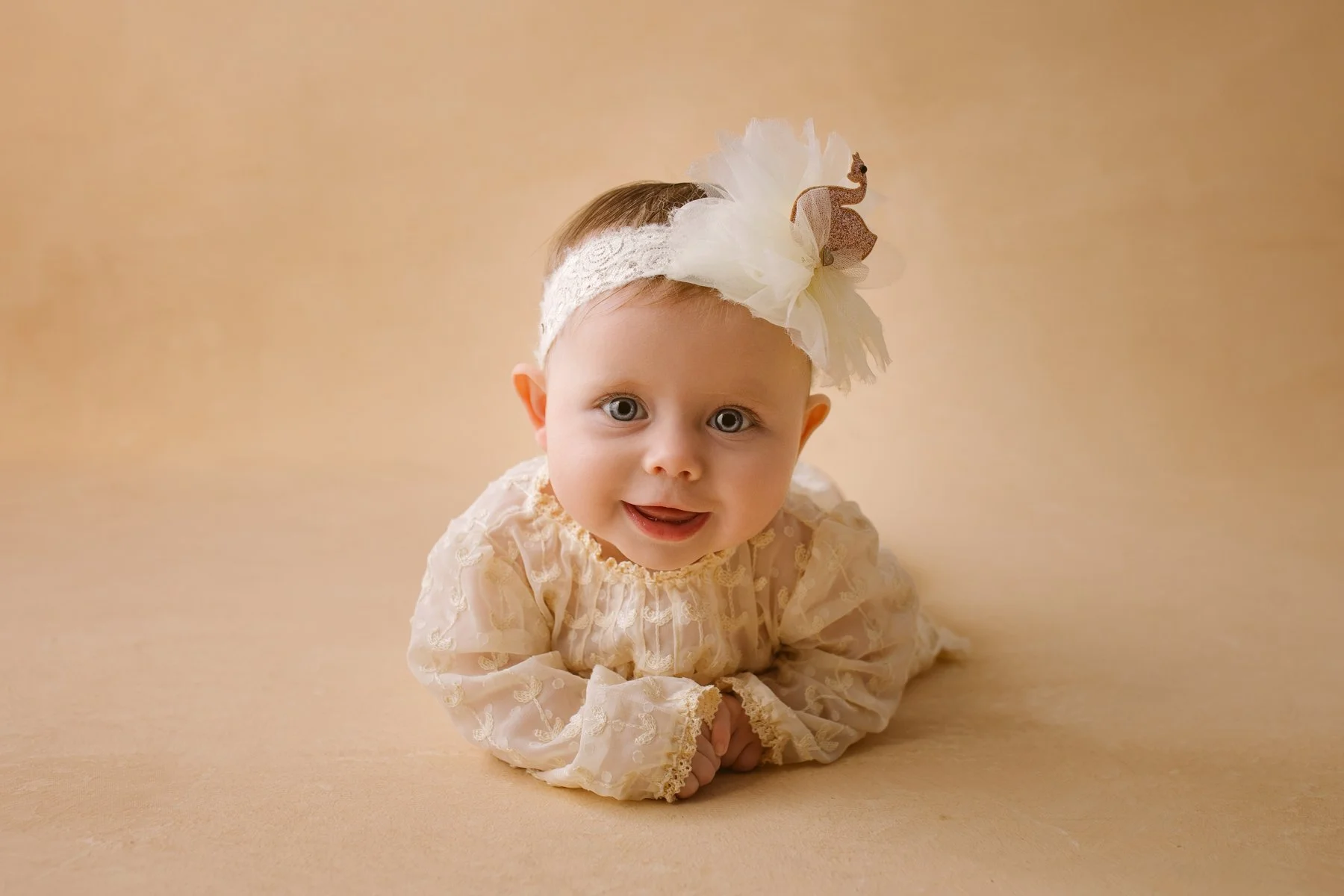 A baby girl with light skin and blue eyes lying on her stomach on a beige surface and background, wearing a cream-colored lace dress and a white headband with a large flower and a small giraffe ornament.