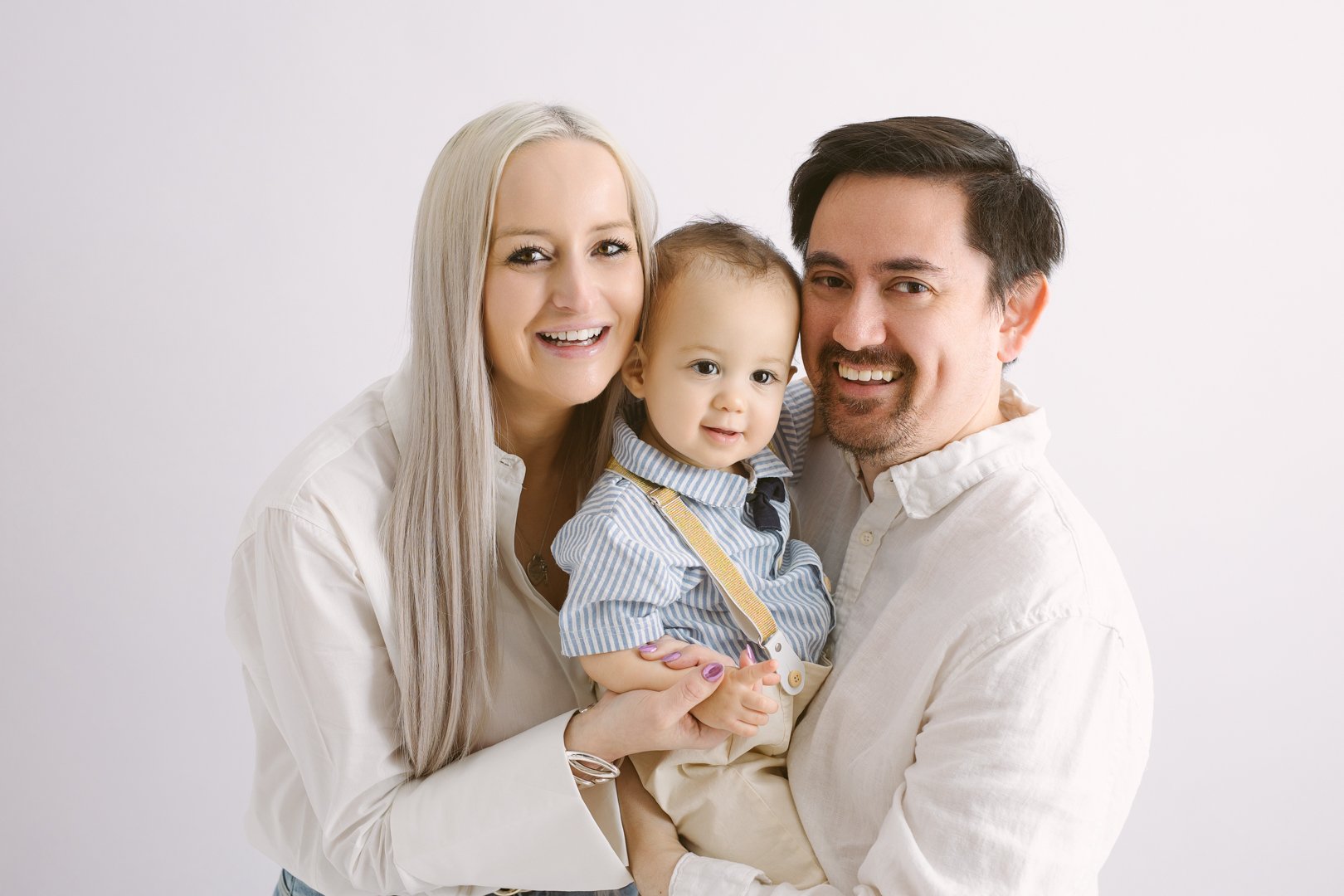 A happy family of three with light skin posing together against a plain white background. The mother with long, straight, platinum blonde hair, wearing a white shirt, holds their young son. The father with dark hair and facial hair, also in a white s