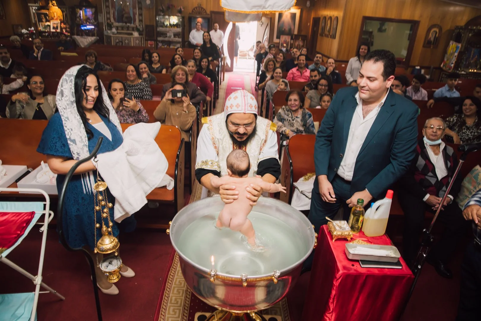 Baby being baptized in a church by a priest while family and friends watch, some taking photos. The scene is joyful with many smiling faces.