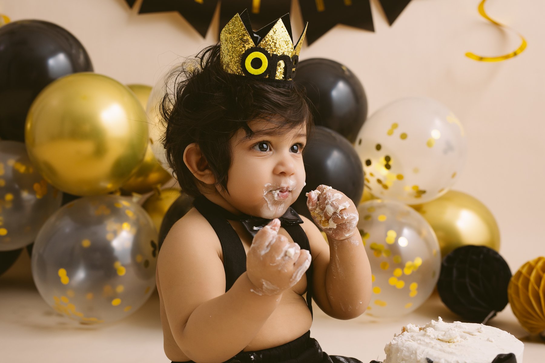 Young child celebrating a birthday with black, gold, and clear balloons, wearing a party hat with 'ONE' on it, and eating cake with frosting.