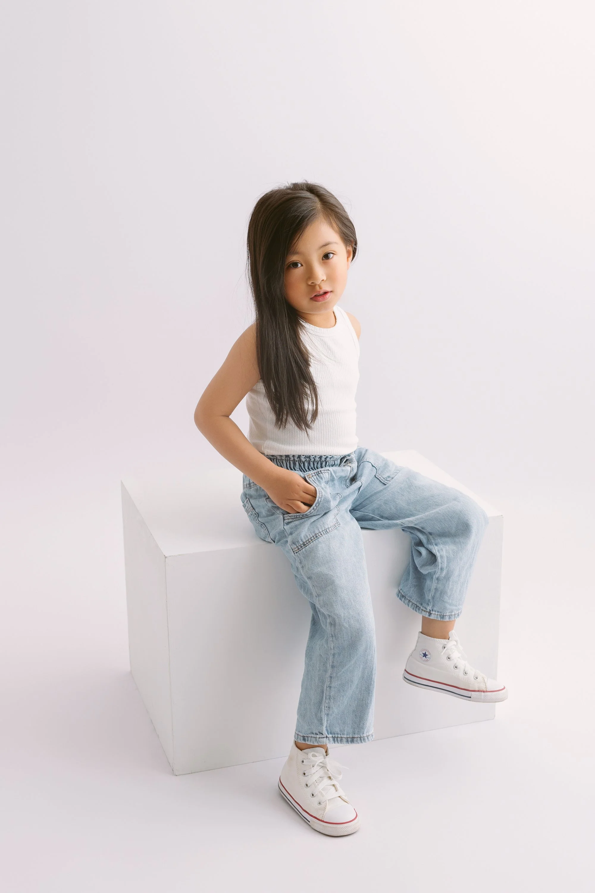 Young girl with long dark hair sitting on a white block against a plain white background, wearing a white sleeveless top, light blue jeans, and white high-top sneakers. She has one hand in her pocket and is looking at the camera.