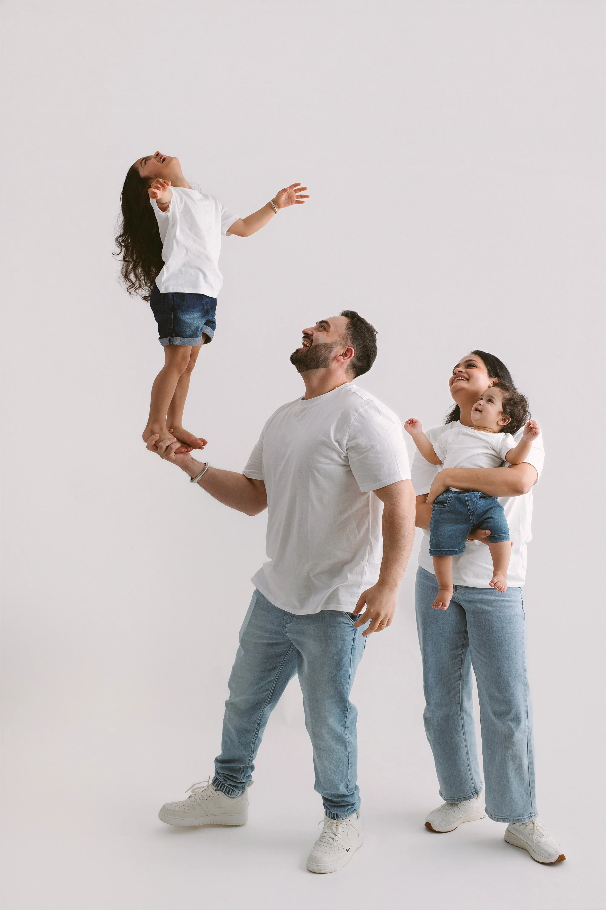 A family of four, with a man and woman holding two young girls, is having fun against a plain white background. The man supports one girl on his hand, who is smiling and laughing, while the woman holds the other girl, both enjoying their time.