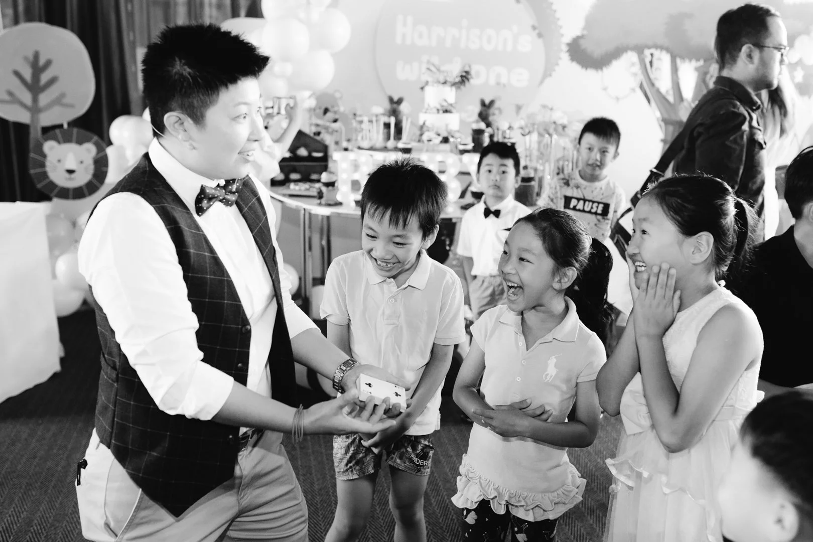 A magician performs a card trick for three children at a birthday party, all smiling and laughing in a decorated party room.