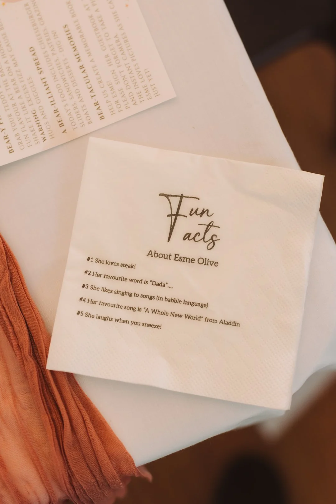 A white paper napkin with the heading 'Fun facts' about Esme Olive, listing her favorite things and amusing quirks, placed on a white tablecloth near some orange fabric and a partially visible printed card.