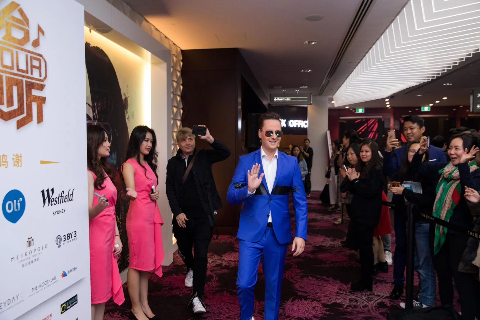 Man in a blue suit and sunglasses waving at fans as he walks through a carpet event surrounded by photographers and fans.