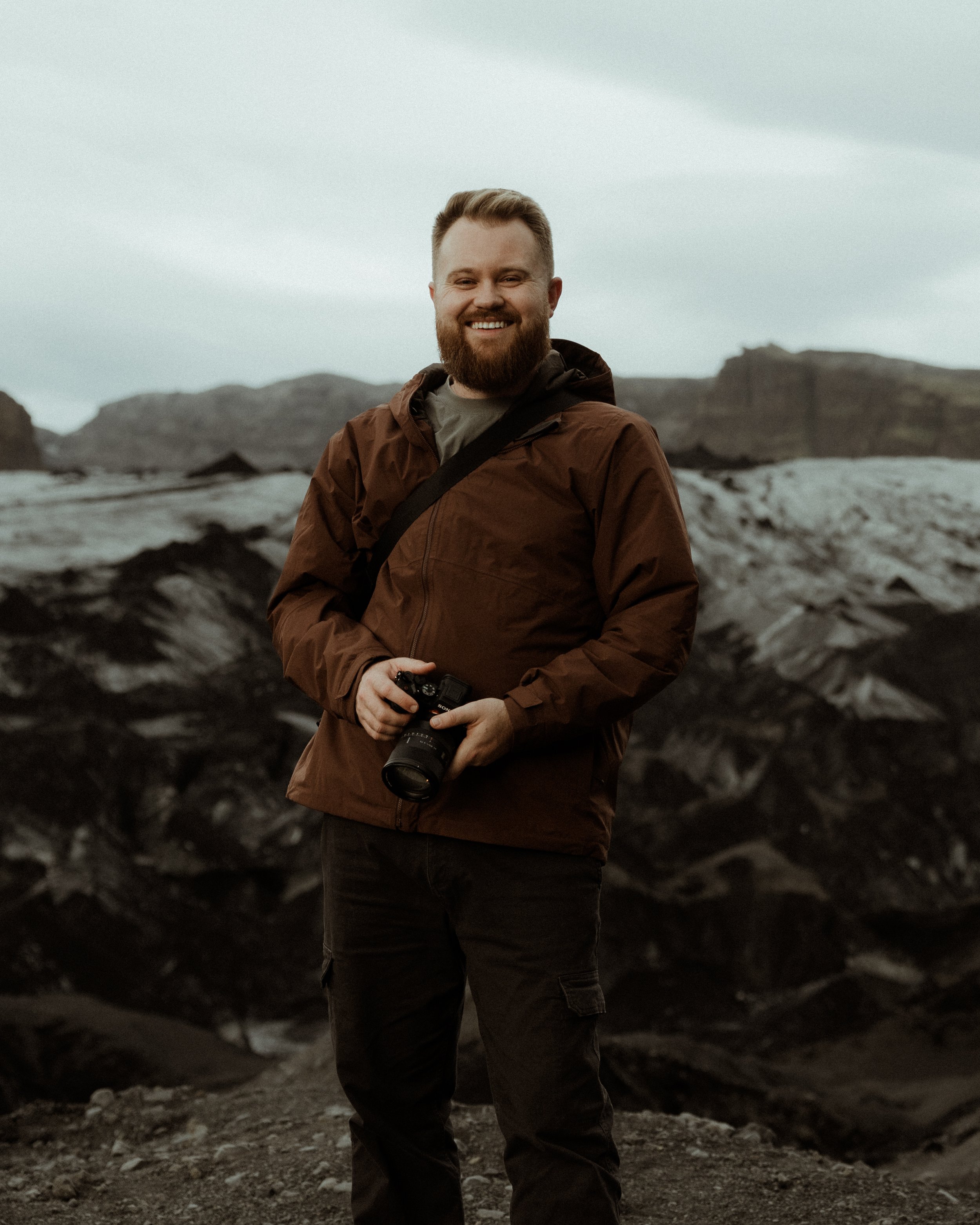 A man with a beard and short hair, wearing a brown jacket and black pants, holding a camera, standing outdoors in a rocky area with mountains and cloudy sky in the background.