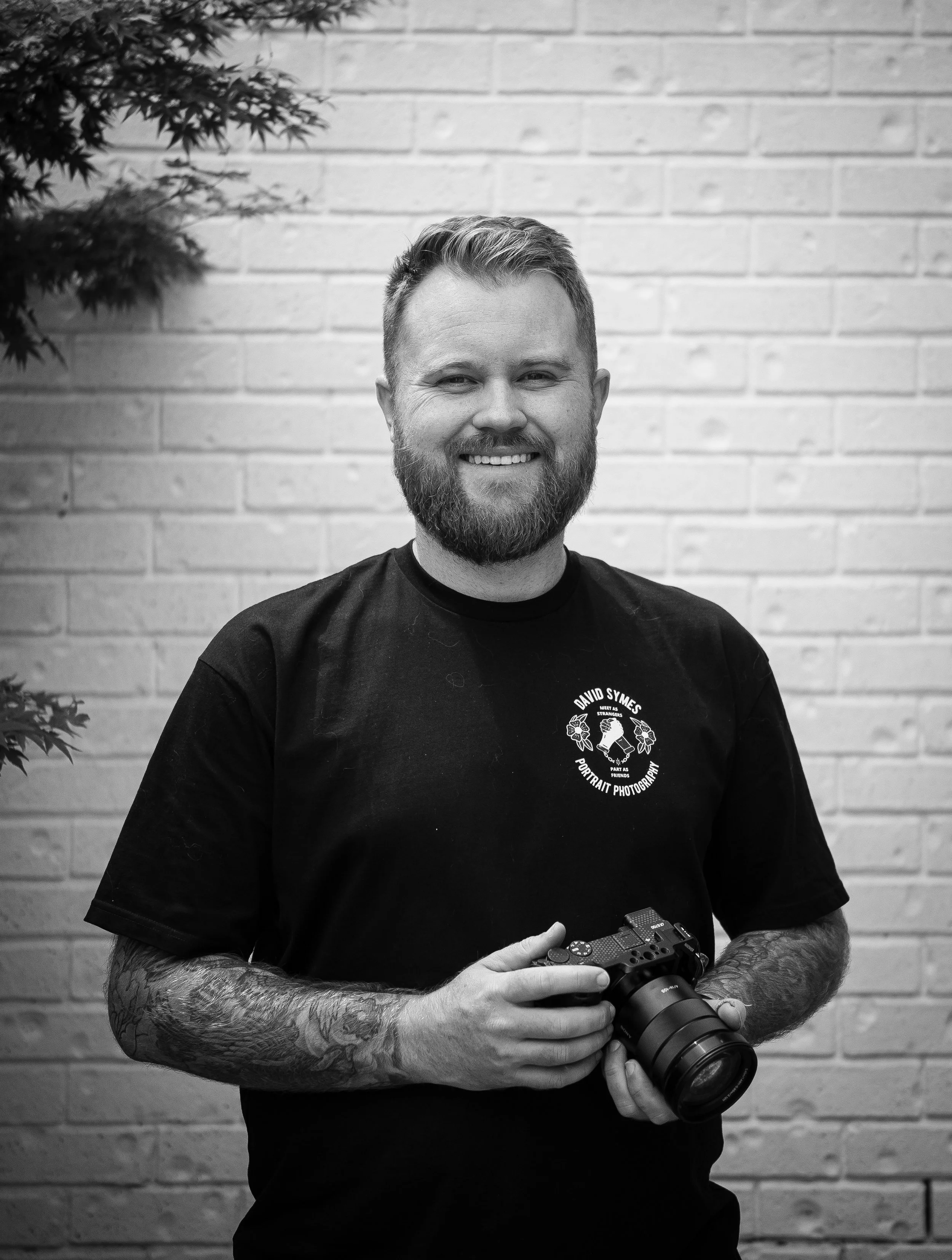 A man with a beard and tattoos on his arms, smiling while holding a camera, standing in front of a brick wall.