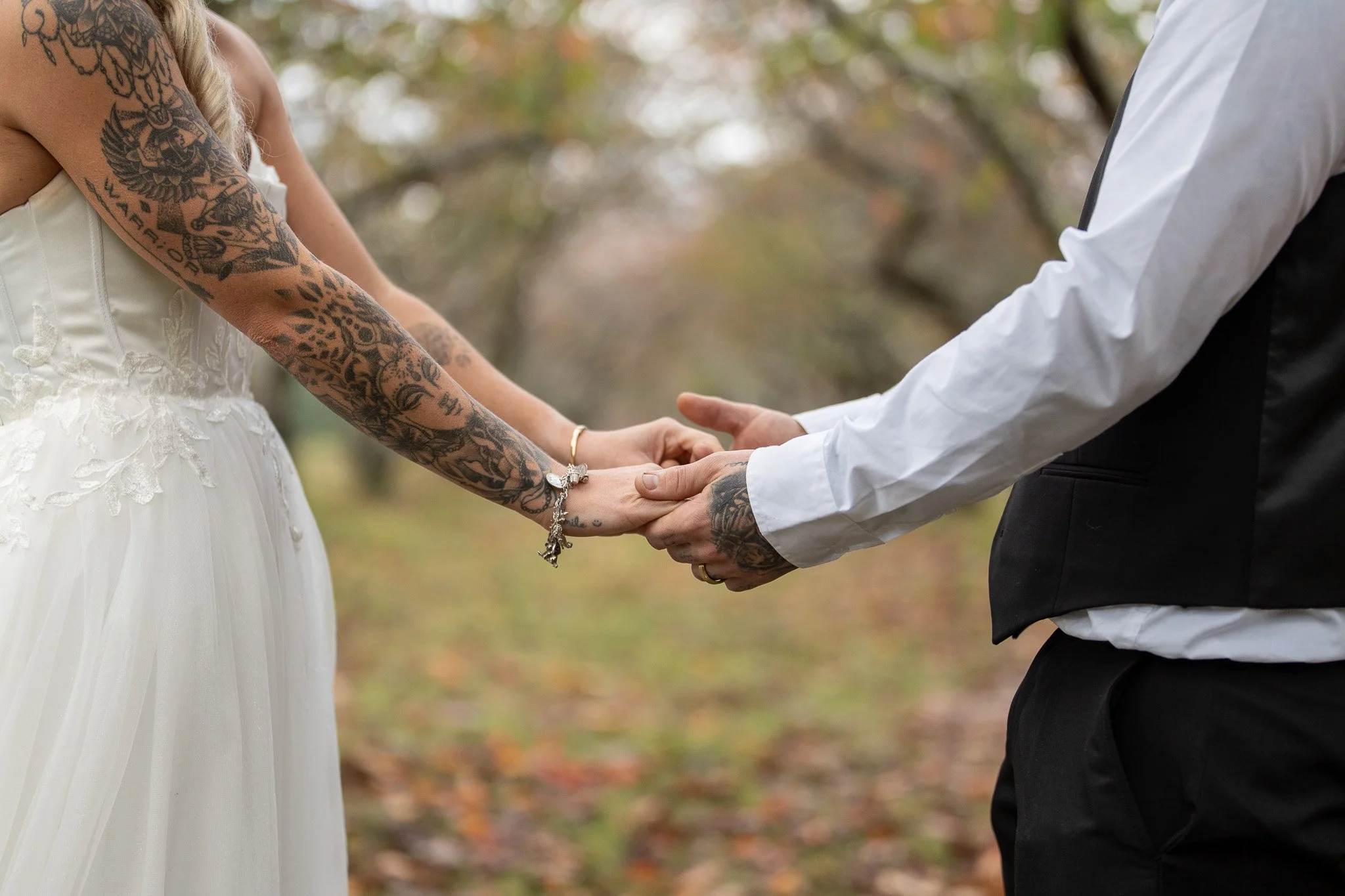 A couple during a wedding ceremony holding hands, with the bride wearing a white wedding dress with lace details and the groom in a white shirt and black vest, outdoors with blurred trees and fallen leaves in the background.