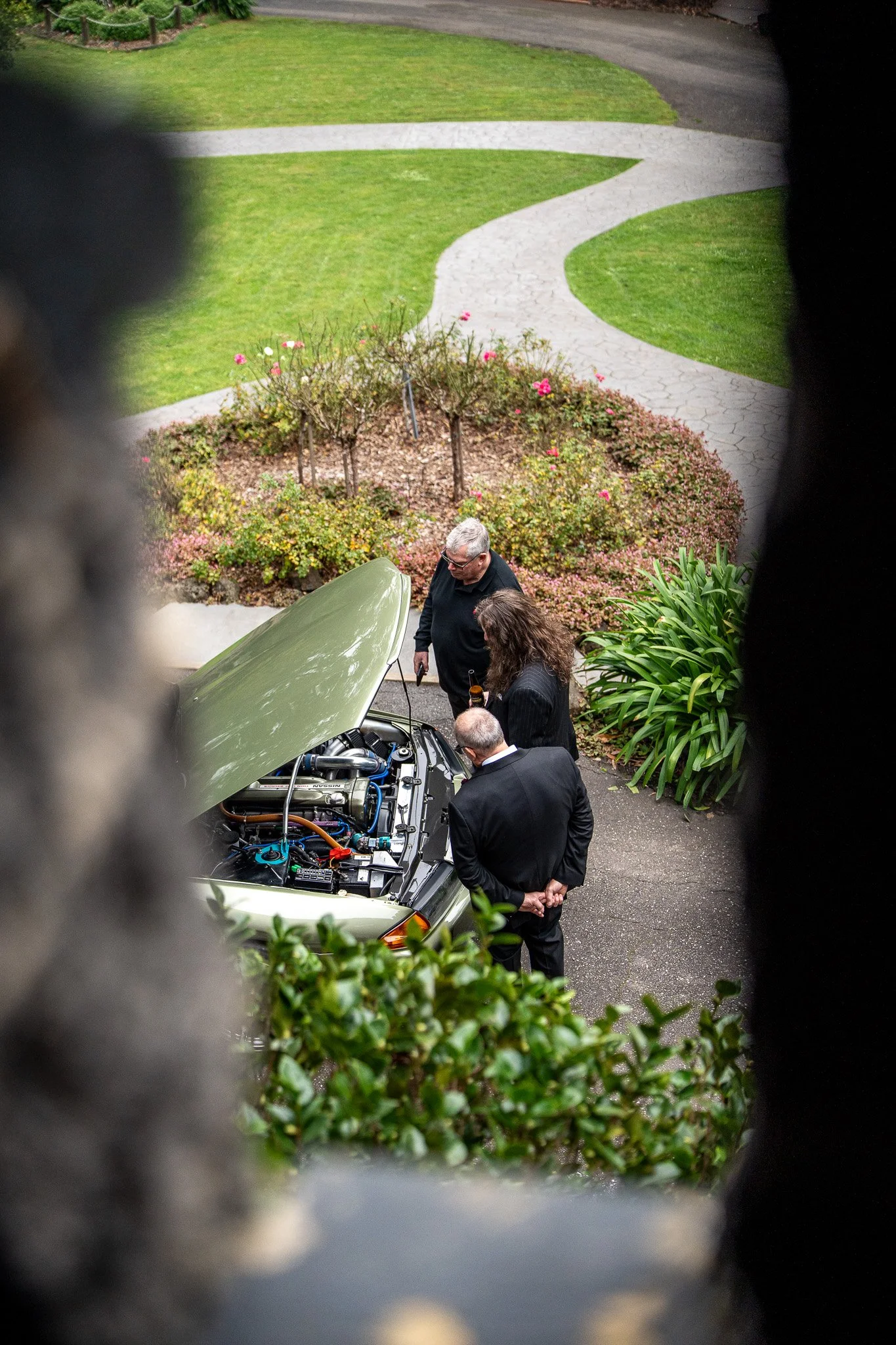 People examining a car engine in a driveway, viewed through a circular opening in a fence or wall, with a well-manicured lawn and brick flower bed in the background.