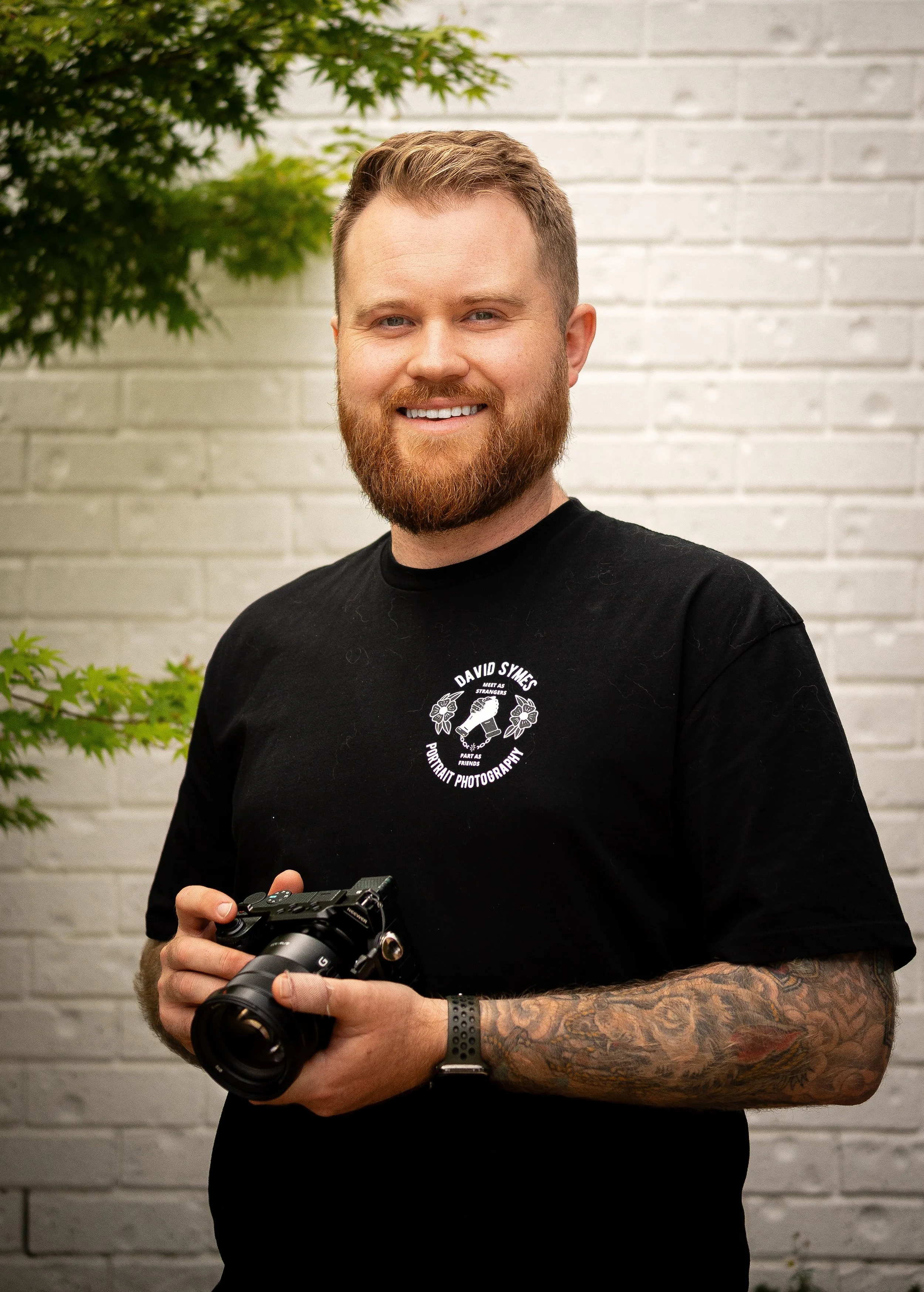 A smiling man with a beard and tattoos, holding a camera, standing outdoors against a white brick wall with a green plant nearby.