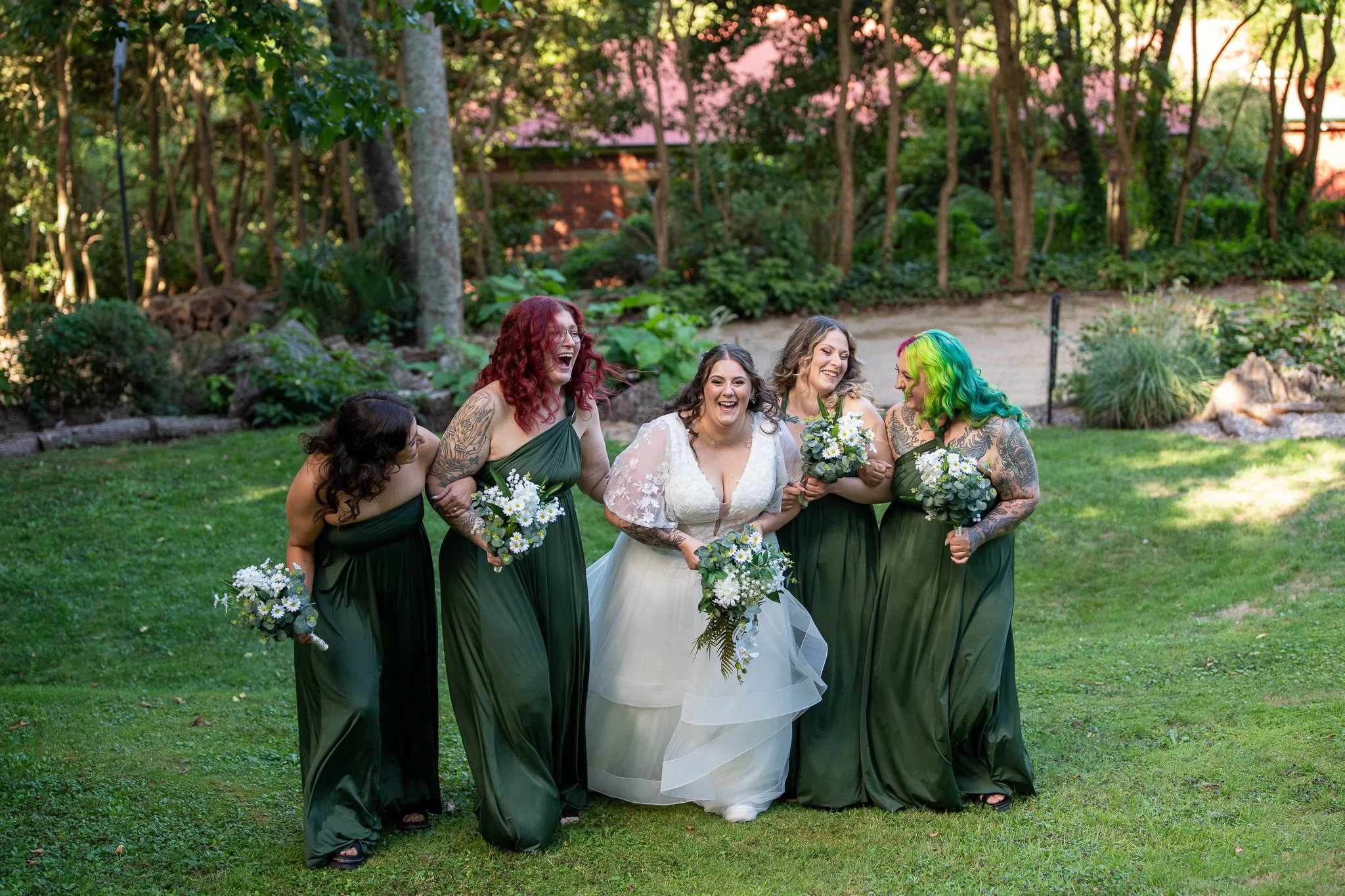 A bride and her four bridesmaids walk and laugh on a grassy area outdoors, surrounded by trees and greenery.