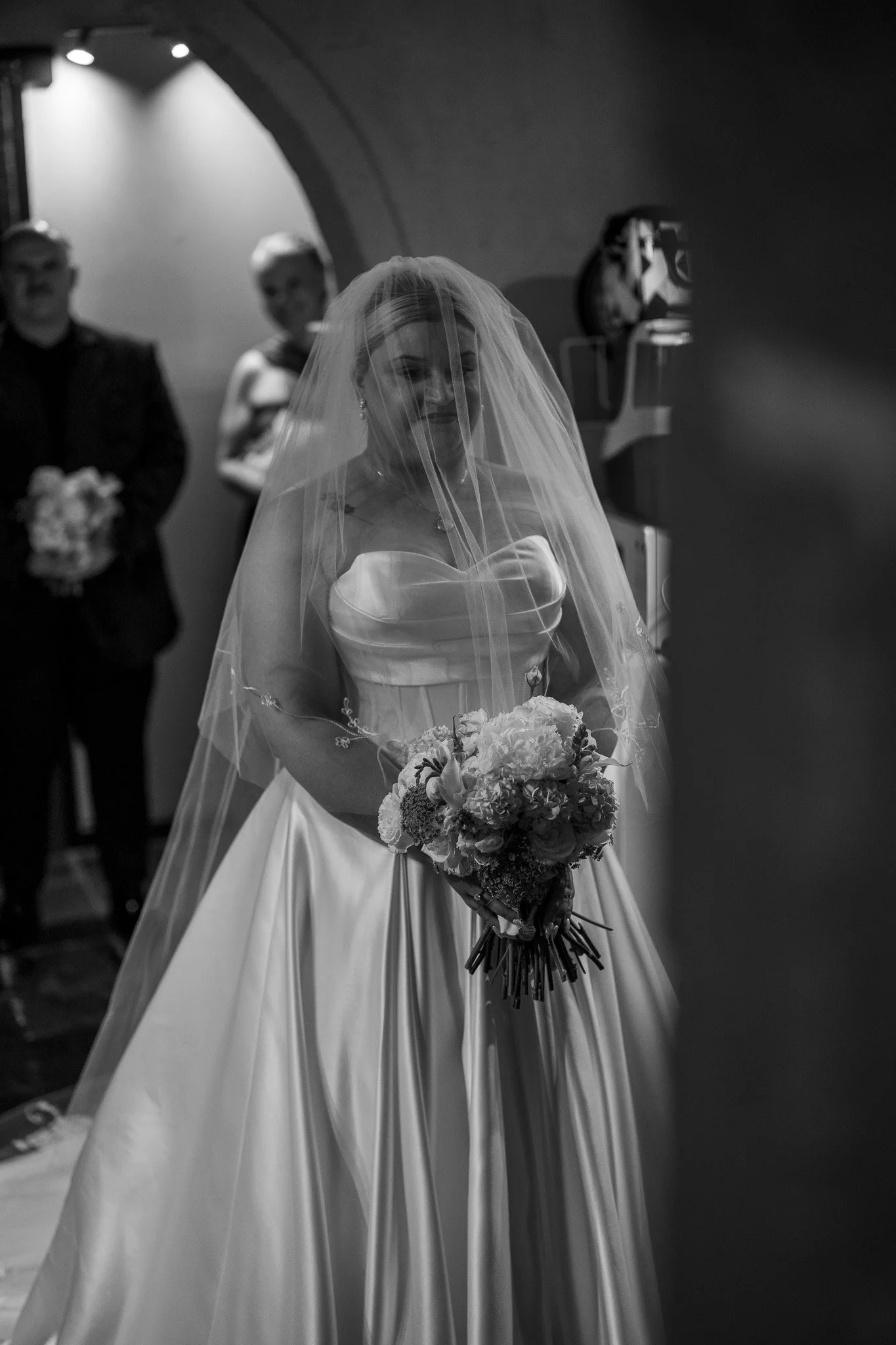 A bride in a wedding gown and veil holding a bouquet of flowers, smiling, with two people in the background and a dark, arched room.