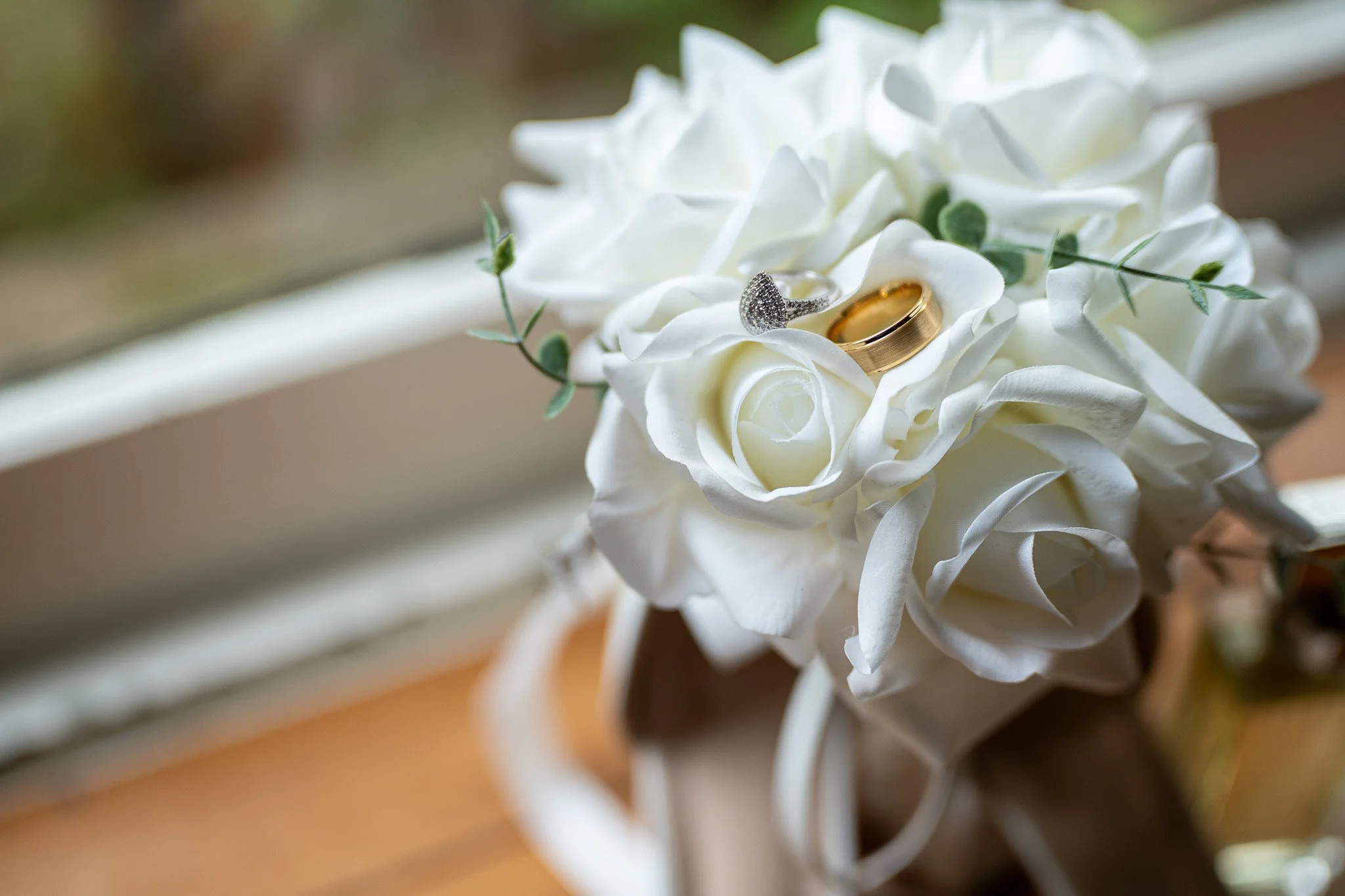 Close-up of wedding rings placed on white artificial flowers with green leaves, sitting in a bouquet or arrangement.