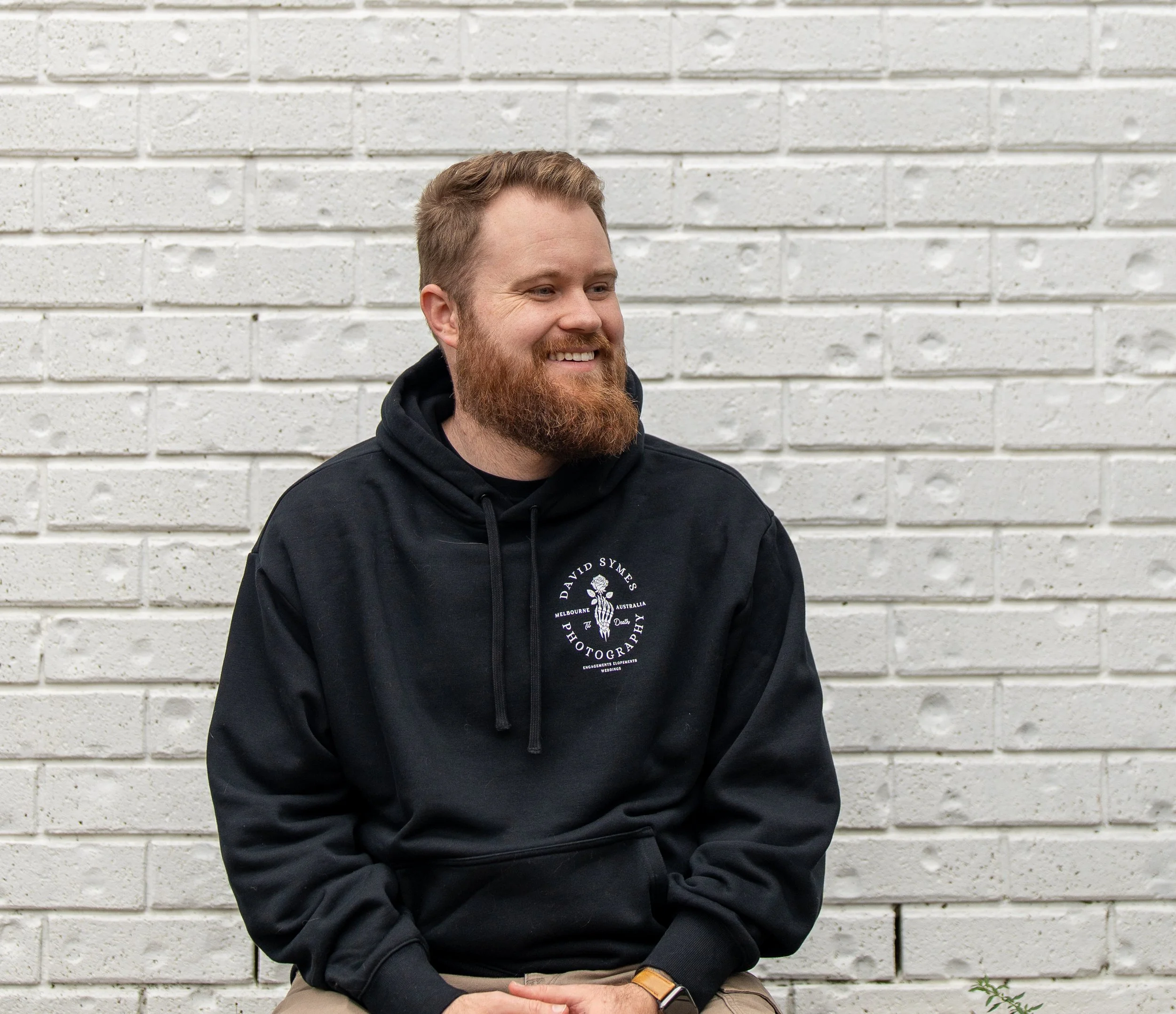 A man with a beard and light brown hair sitting against a white brick wall, wearing a black hoodie with white text and a graphic on it.