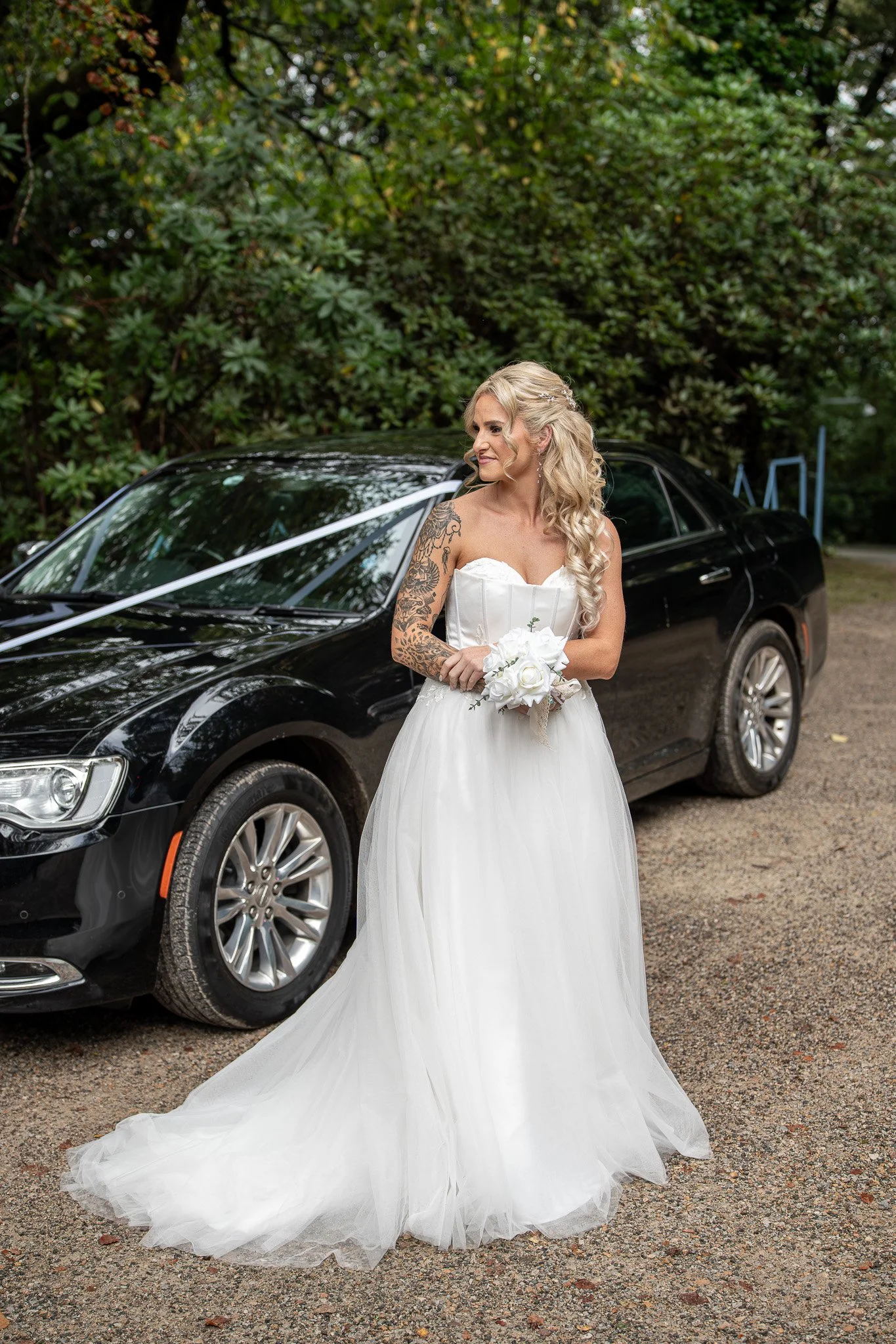 A woman in a white wedding gown holding a small bouquet of white roses stands outdoors next to a black car decorated with white ribbons. She has long blonde curly hair and tattoos on her right arm, and is looking to her left with a slight smile. The 
