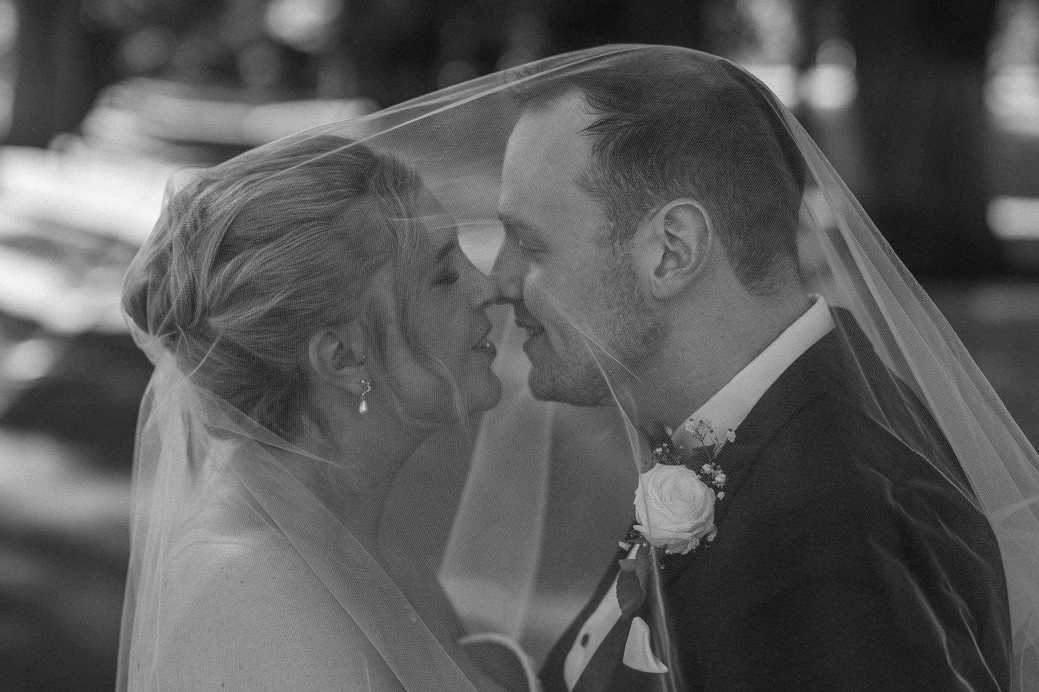 A bride and groom, facing each other with noses touching, under a veil on their wedding day.
