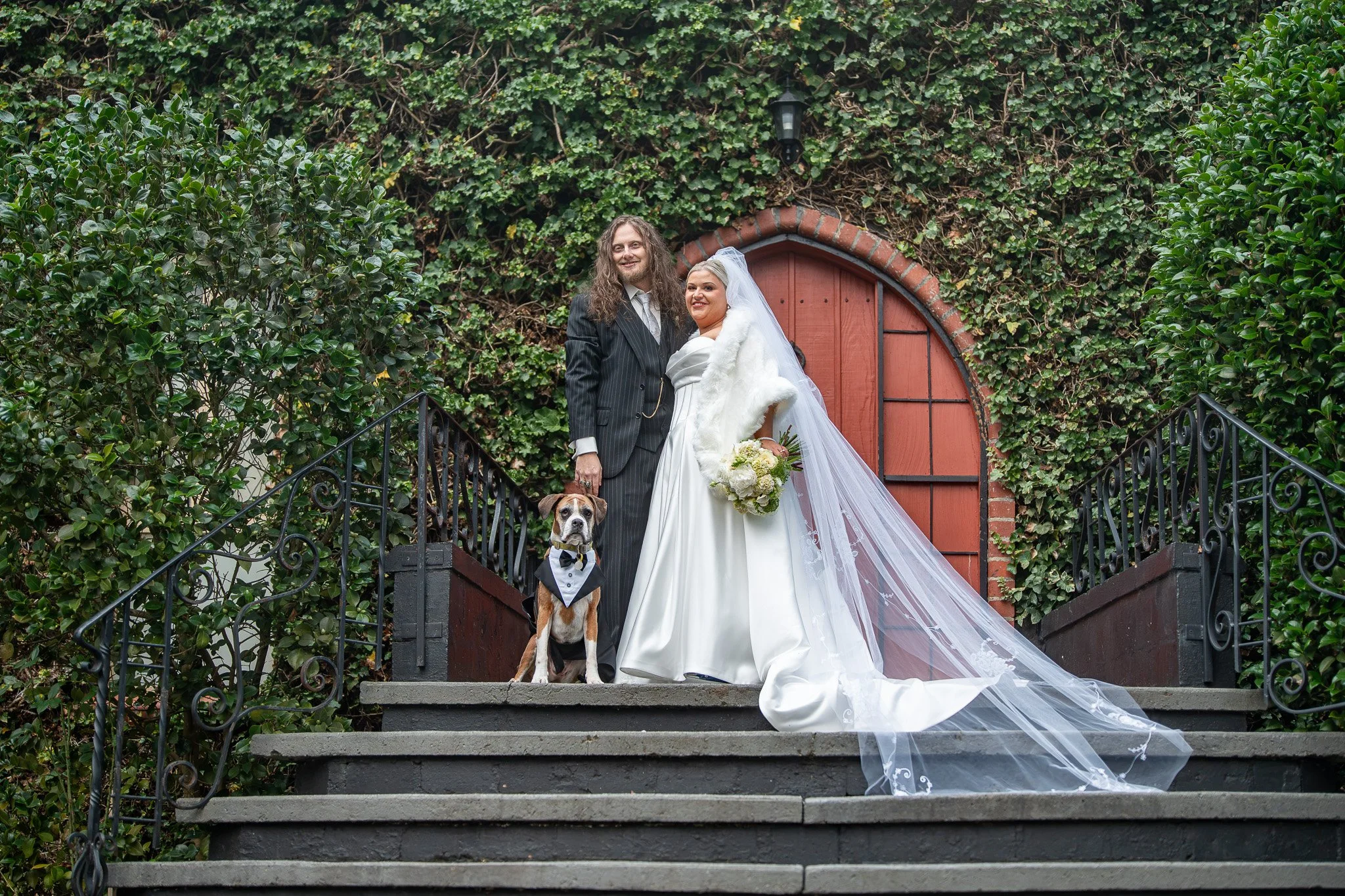 A newlywed couple dressed in wedding attire standing on outdoor stairs with a dog, surrounded by greenery and a red arched gate in the background.