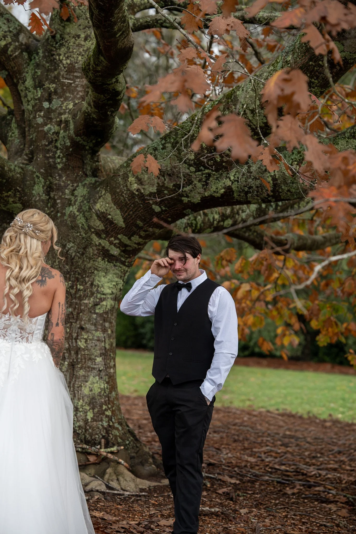 A bride with long blonde hair and tattoos on her shoulder faces a groom with dark hair, wearing a tuxedo and holding his eye, standing under a large moss-covered tree with autumn leaves in the background.