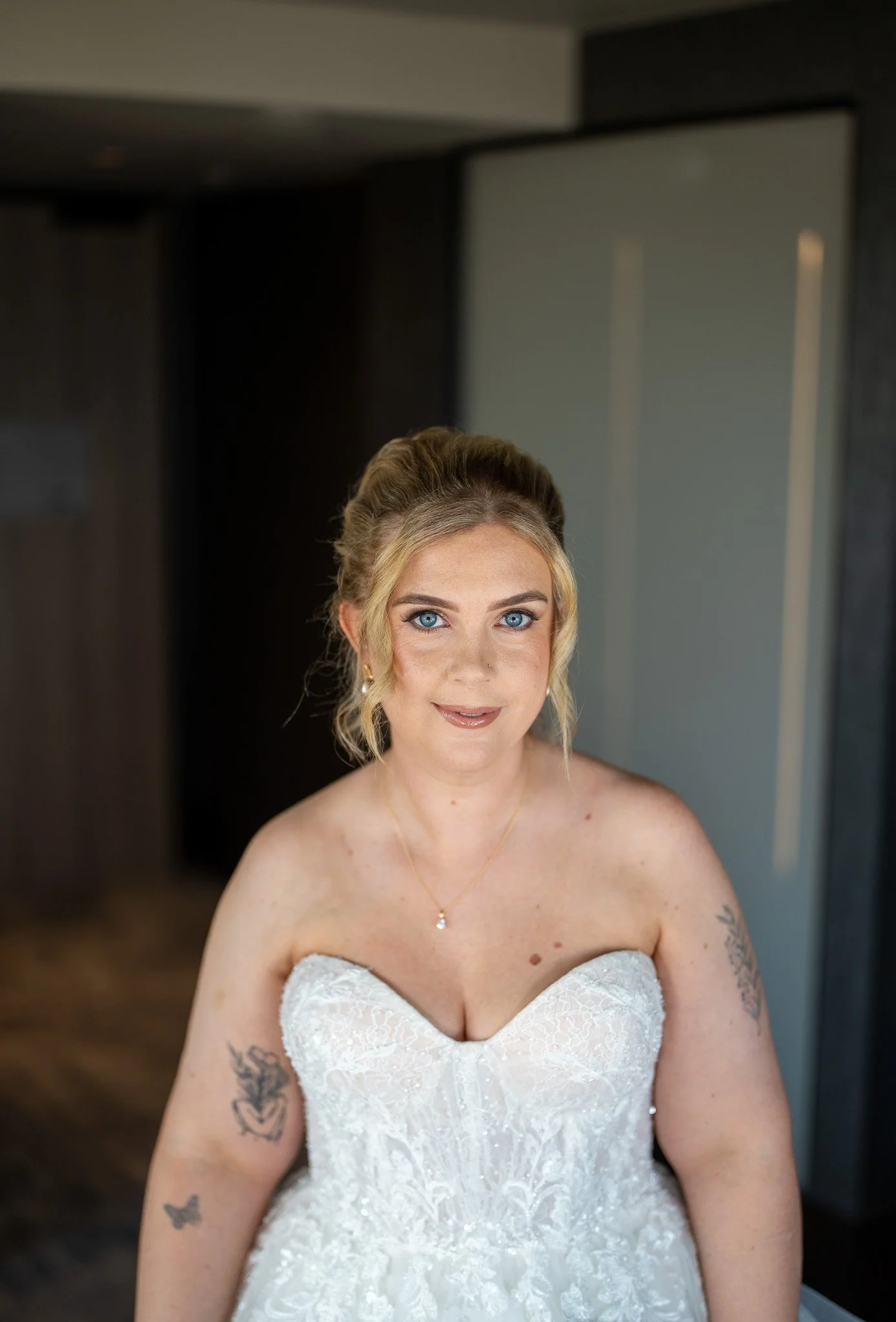 A woman with blonde hair and blue eyes wearing a white strapless lace wedding dress and a gold necklace, standing indoors.