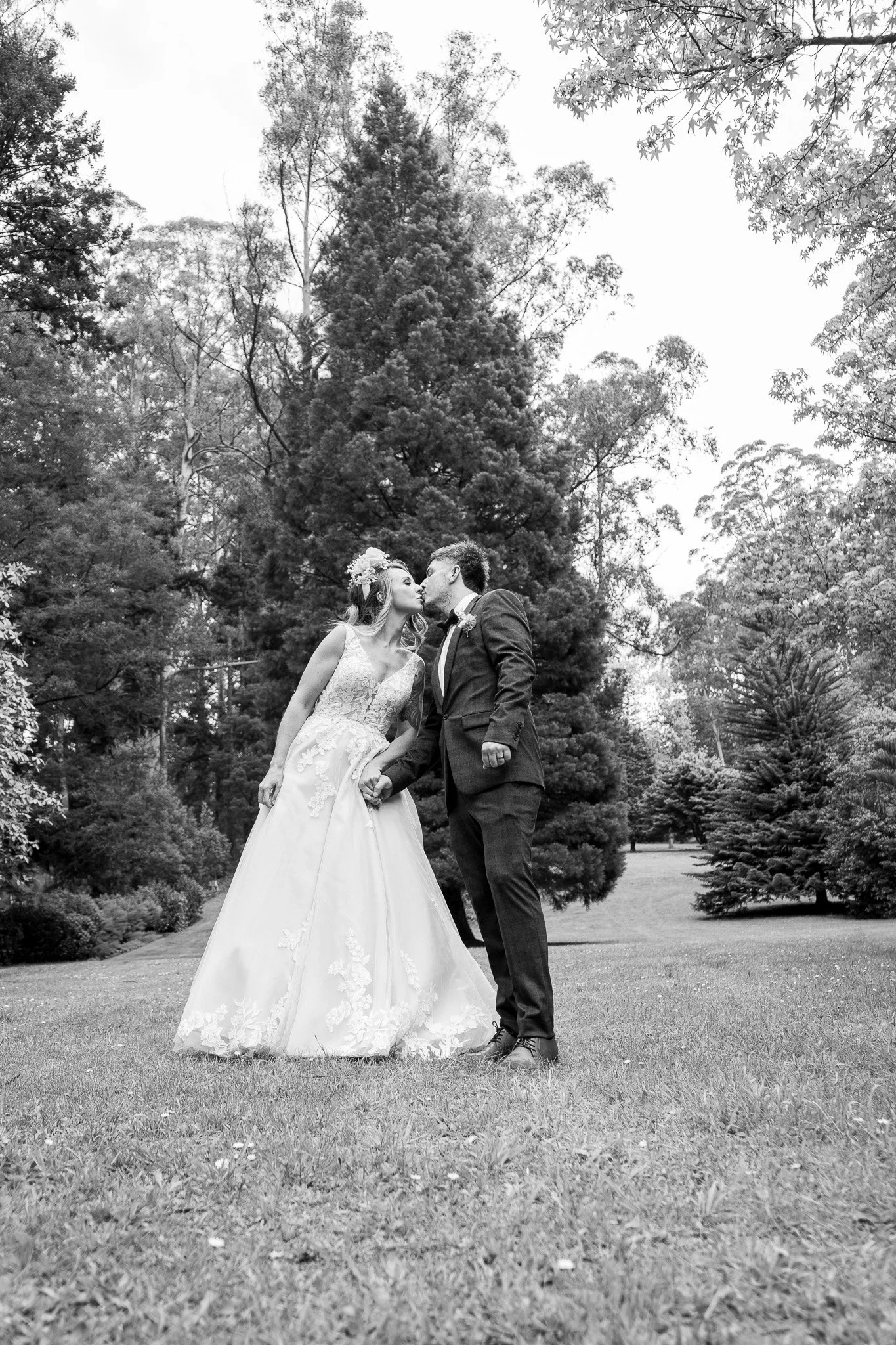 Black and white photo of a bride and groom kissing outdoors, with trees and grass in the background.