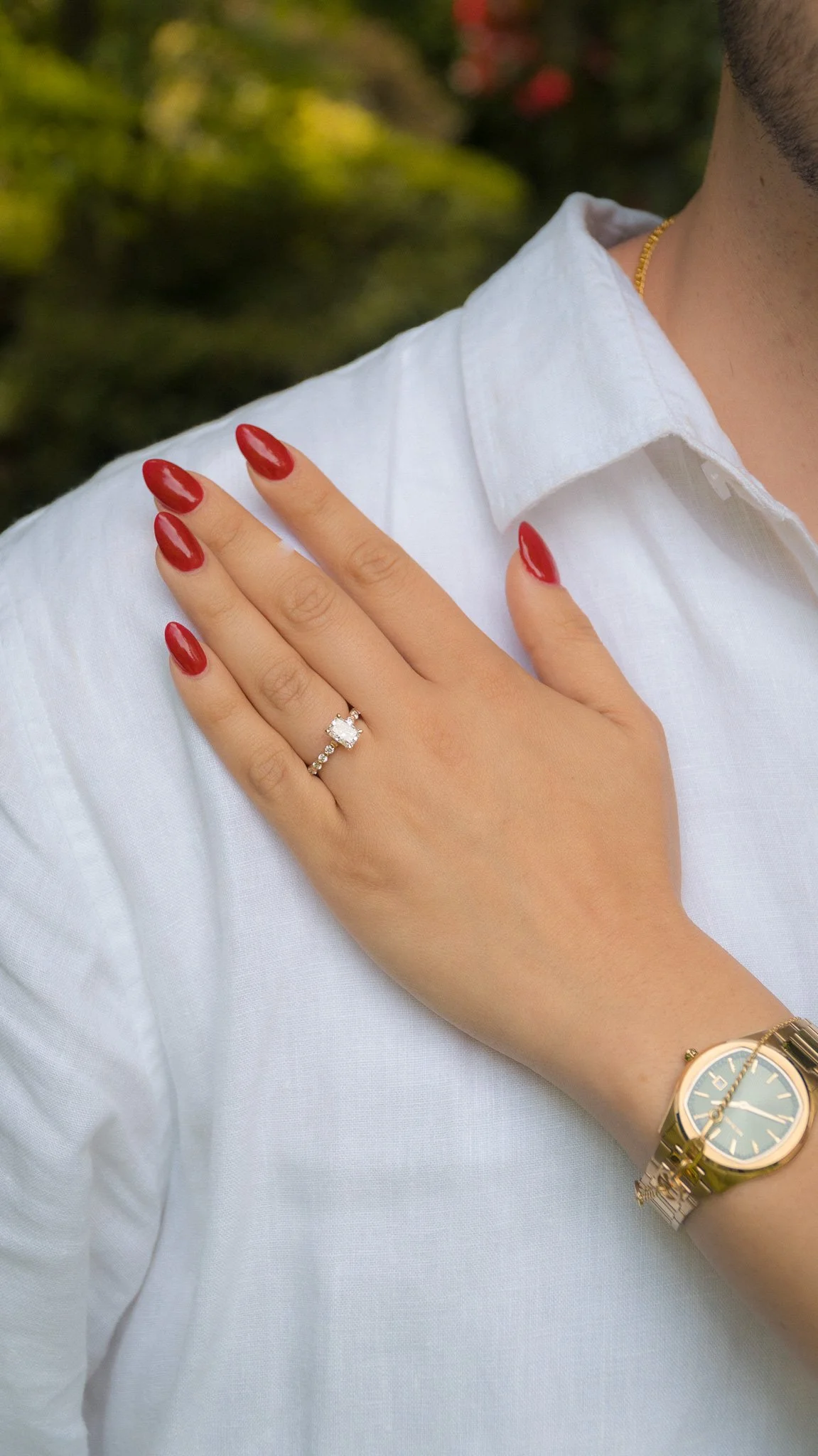 A woman's hand with red polished nails, wearing an engagement ring with a large square-cut diamond and a wedding band with small diamonds, resting on a man's shoulder. The man is wearing a white shirt, a gold watch, and a gold chain. There is a blurr
