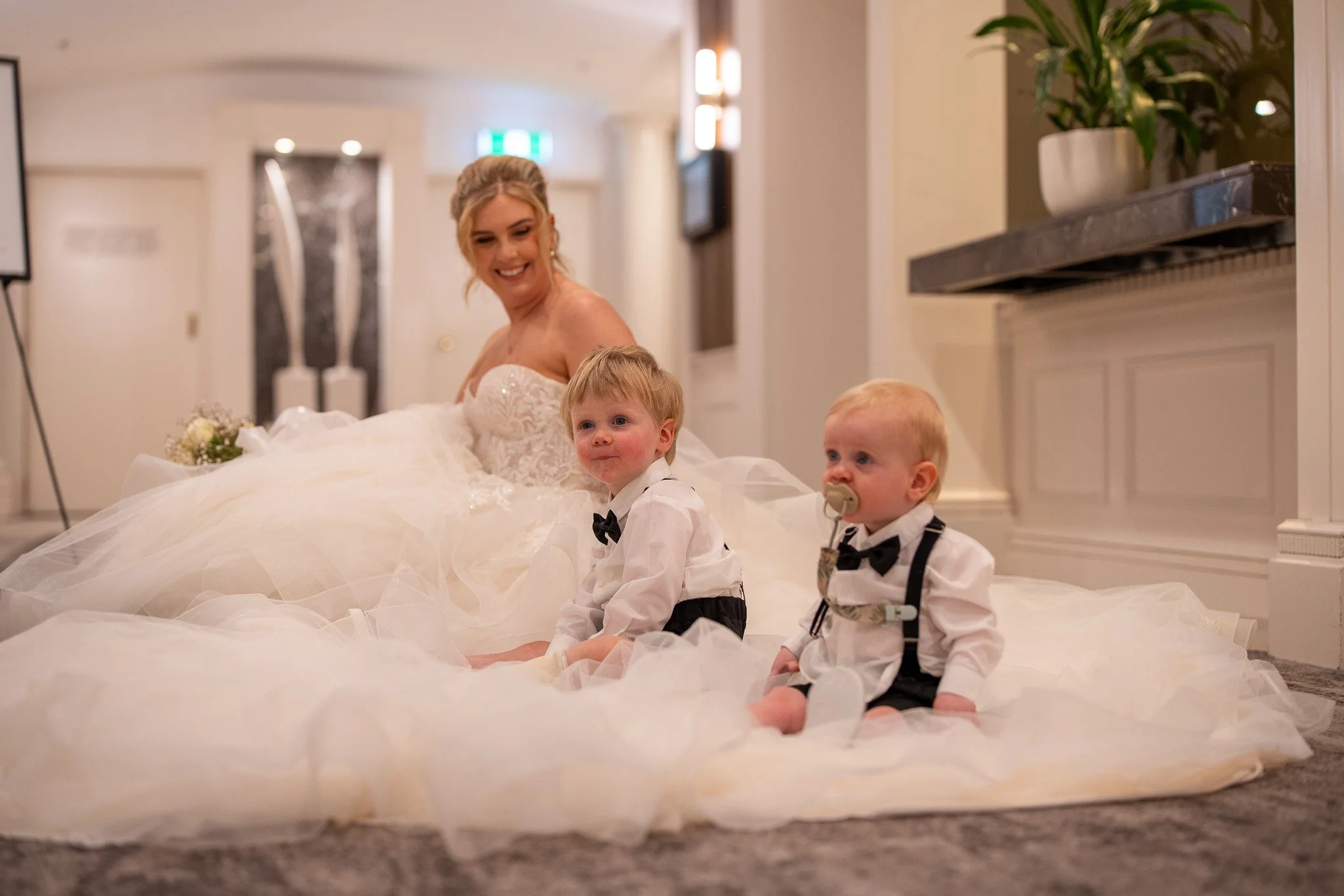 A smiling bride in a wedding dress sitting on the floor with two young boys dressed in tuxedos, one with a pacifier, in an indoor setting.