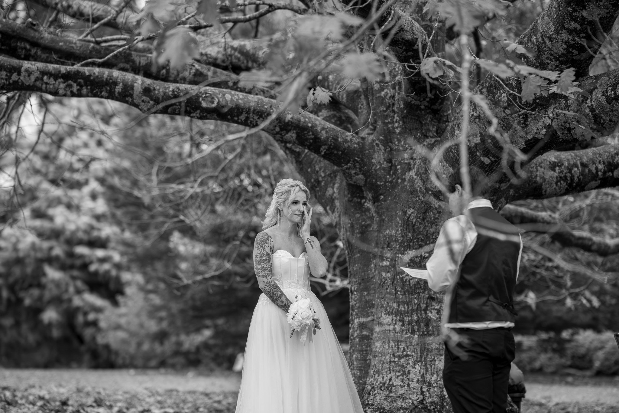 A bride in a white strapless wedding dress with tattoos on her arms, holding a bouquet of flowers, standing outdoors beside a person in formal attire, underneath a large tree.