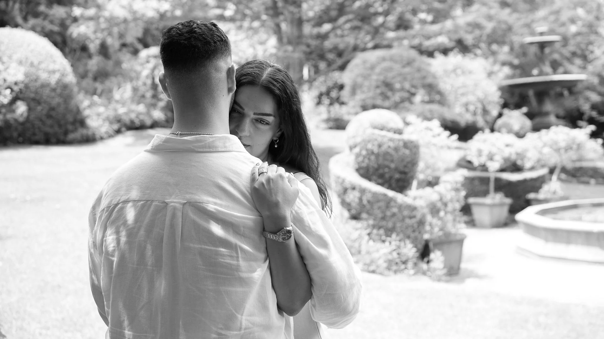 A couple embraces in a garden, with the woman resting her head on the man's shoulder, surrounded by well-manicured bushes and trees.