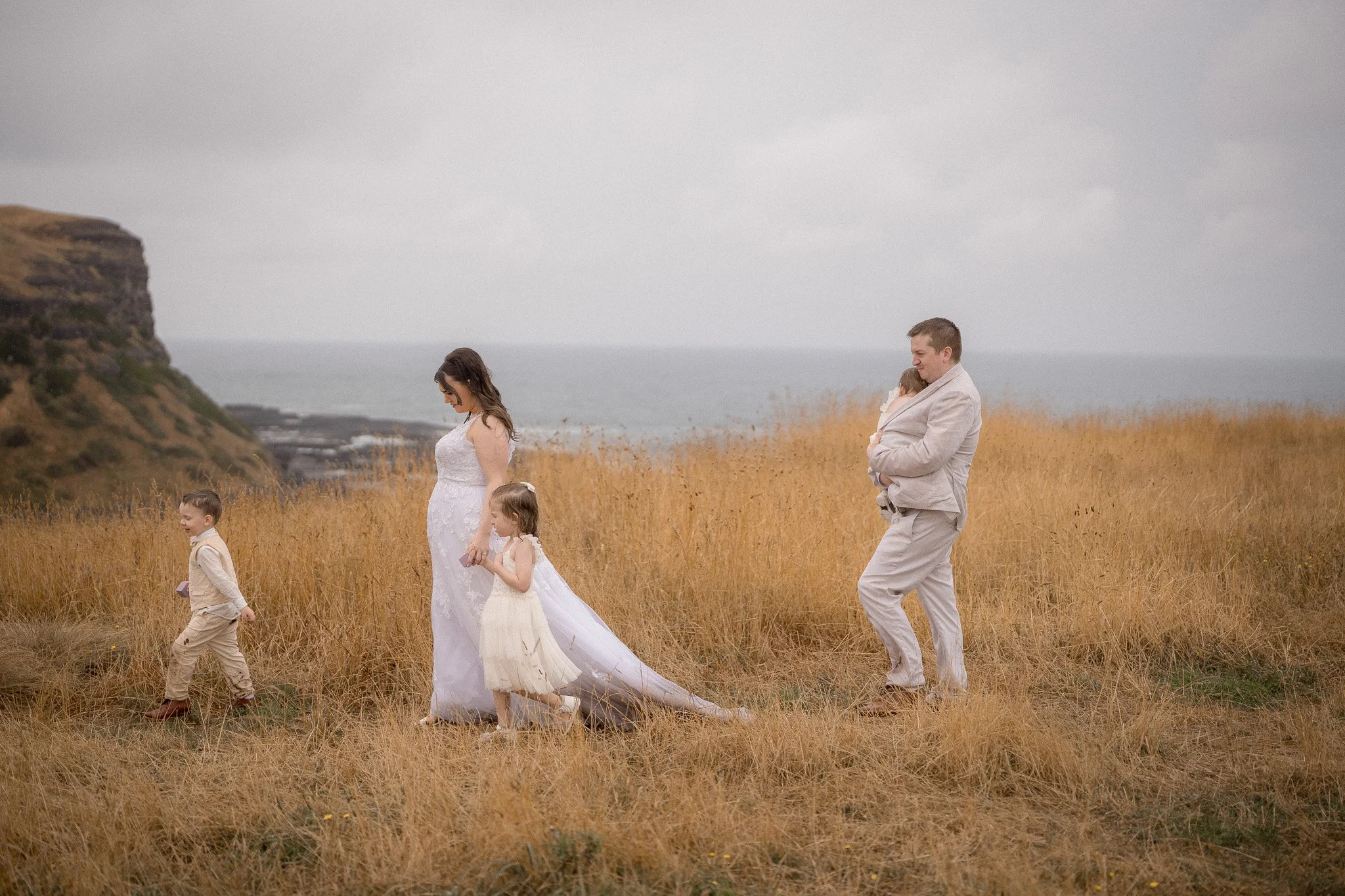 A family in wedding attire walking on a grassy field near the coast, with cliffs and the ocean in the background. The woman is in a wedding dress, two children in formal clothing walk nearby, and a man in a light-colored suit holds a smaller child.
