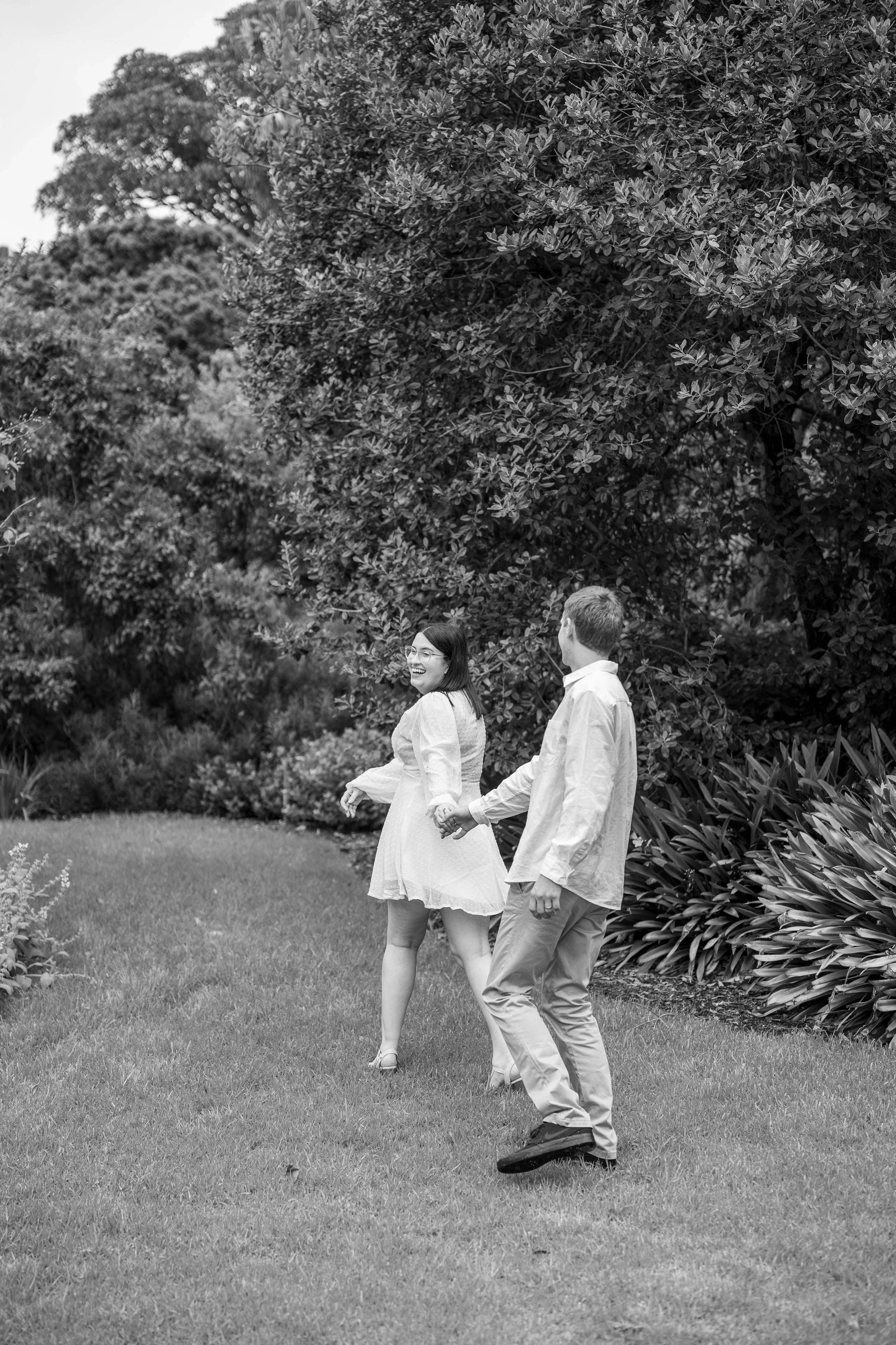 A smiling woman and man walking hand in hand in a garden with lush foliage in black and white.