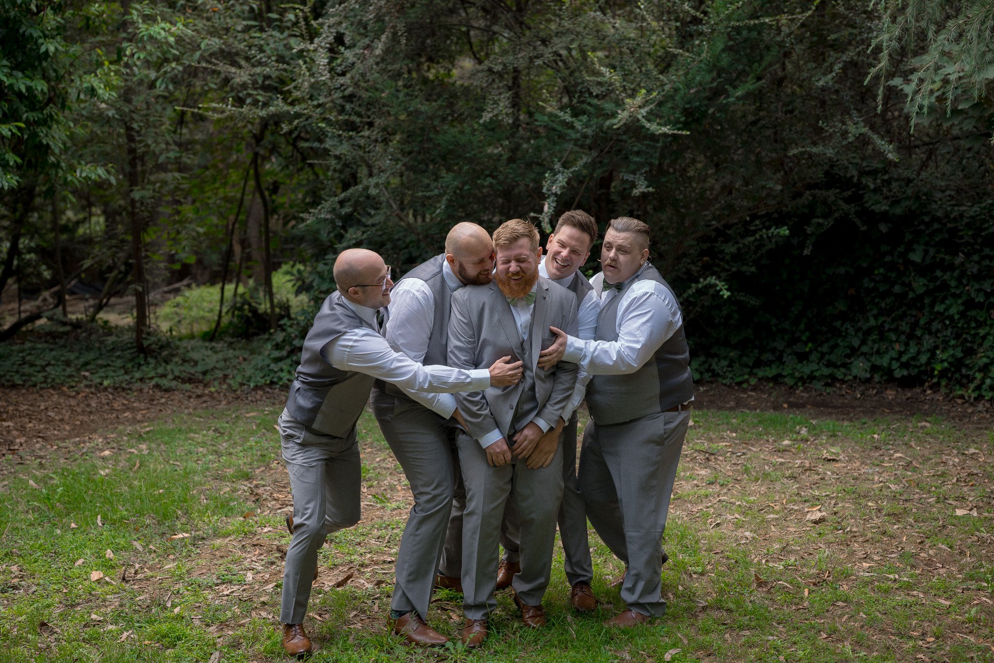 A group of five men in suits and vests are playfully pretending to fight or wrestle in a wooded outdoor area.