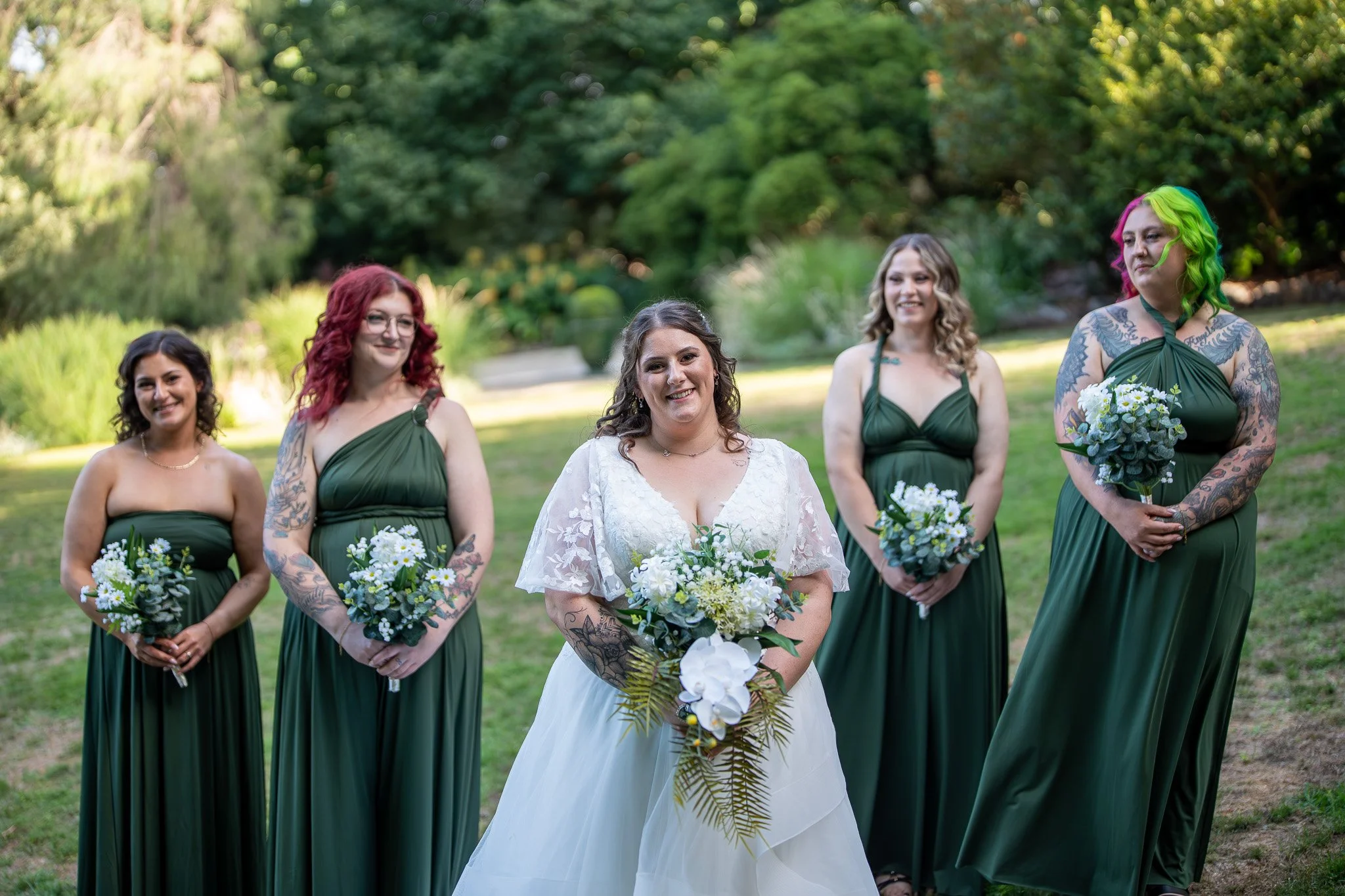 Bride in white dress holding bouquet of flowers standing with bridesmaids in dark green dresses holding bouquets outdoors in a garden