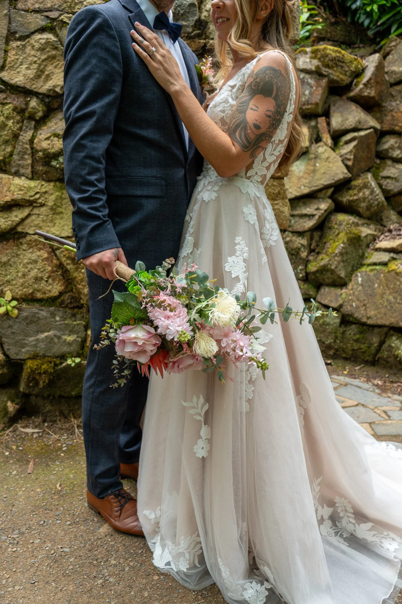 A bride and groom embrace outdoors, with the bride showing a tattoo on her arm, holding a bouquet of pink and white flowers, against a stone wall background.