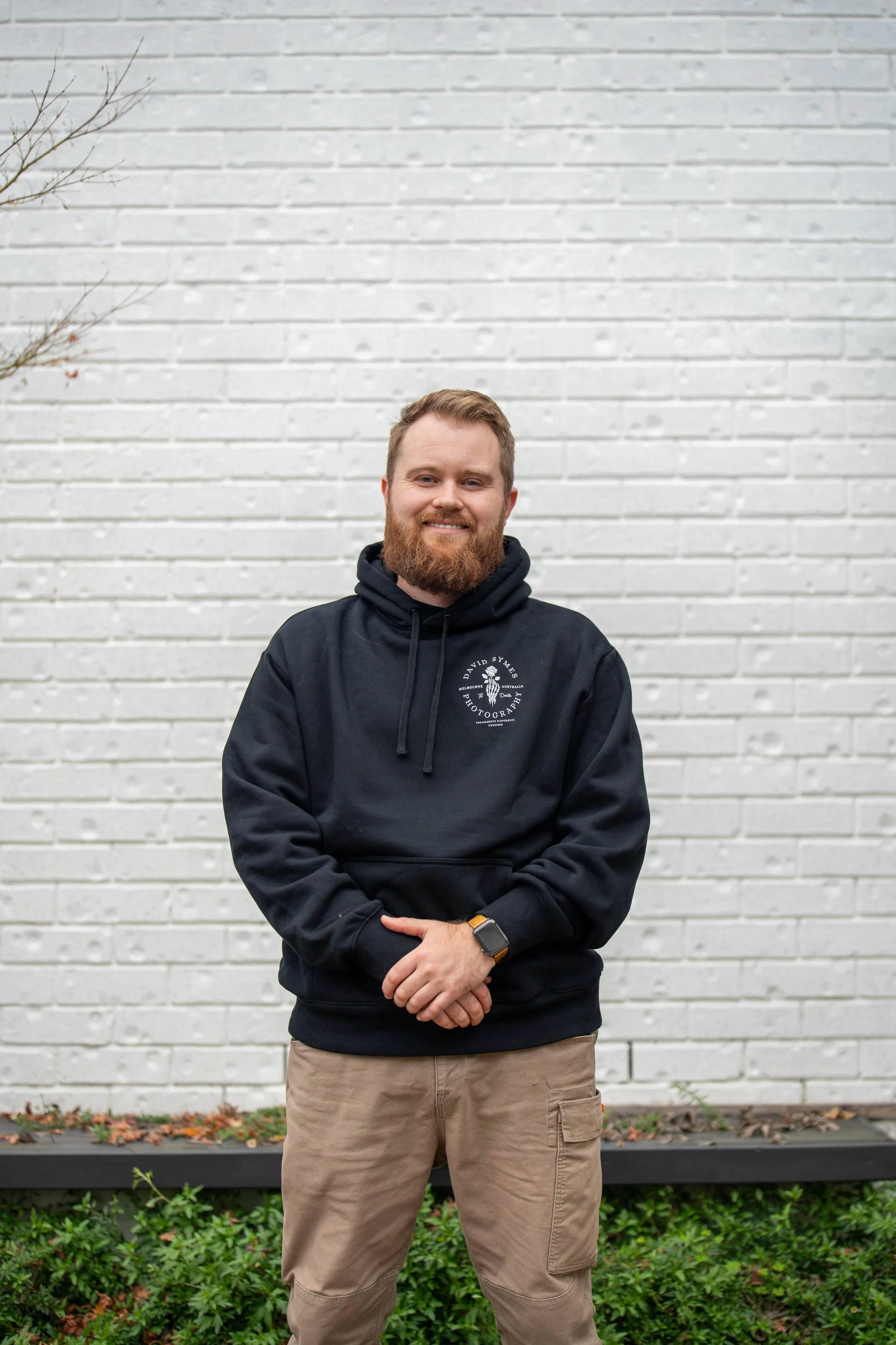 A man with a beard and light brown hair standing outdoors in front of a white brick wall, wearing a black hoodie and tan cargo pants.