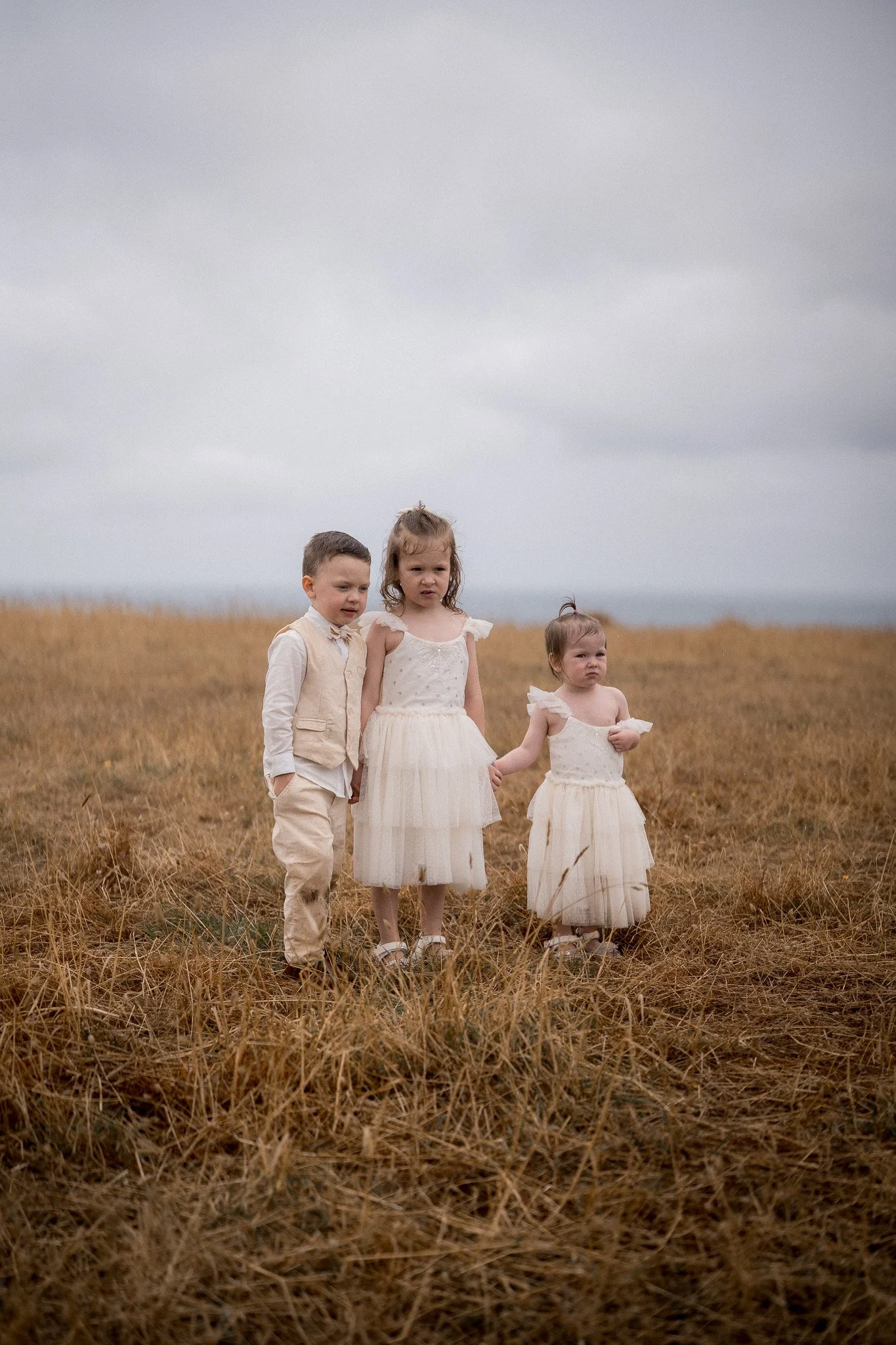 Three young children, two girls and a boy, standing in a dry, grassy field under a cloudy sky, dressed in light-colored, vintage-style clothing.