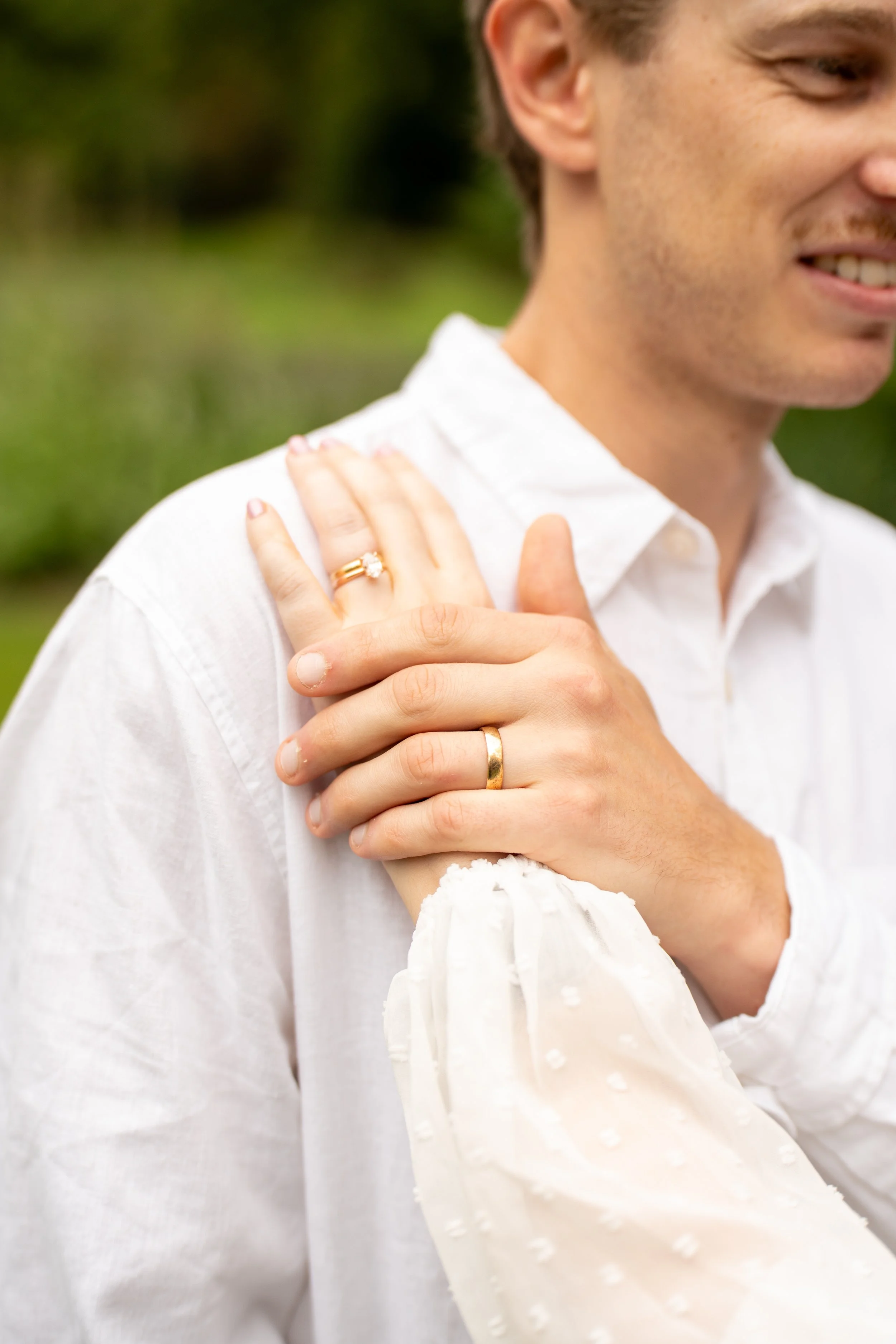 A close-up of two hands, one with a diamond ring, on a man's shoulder outdoors, with a blurred natural background.