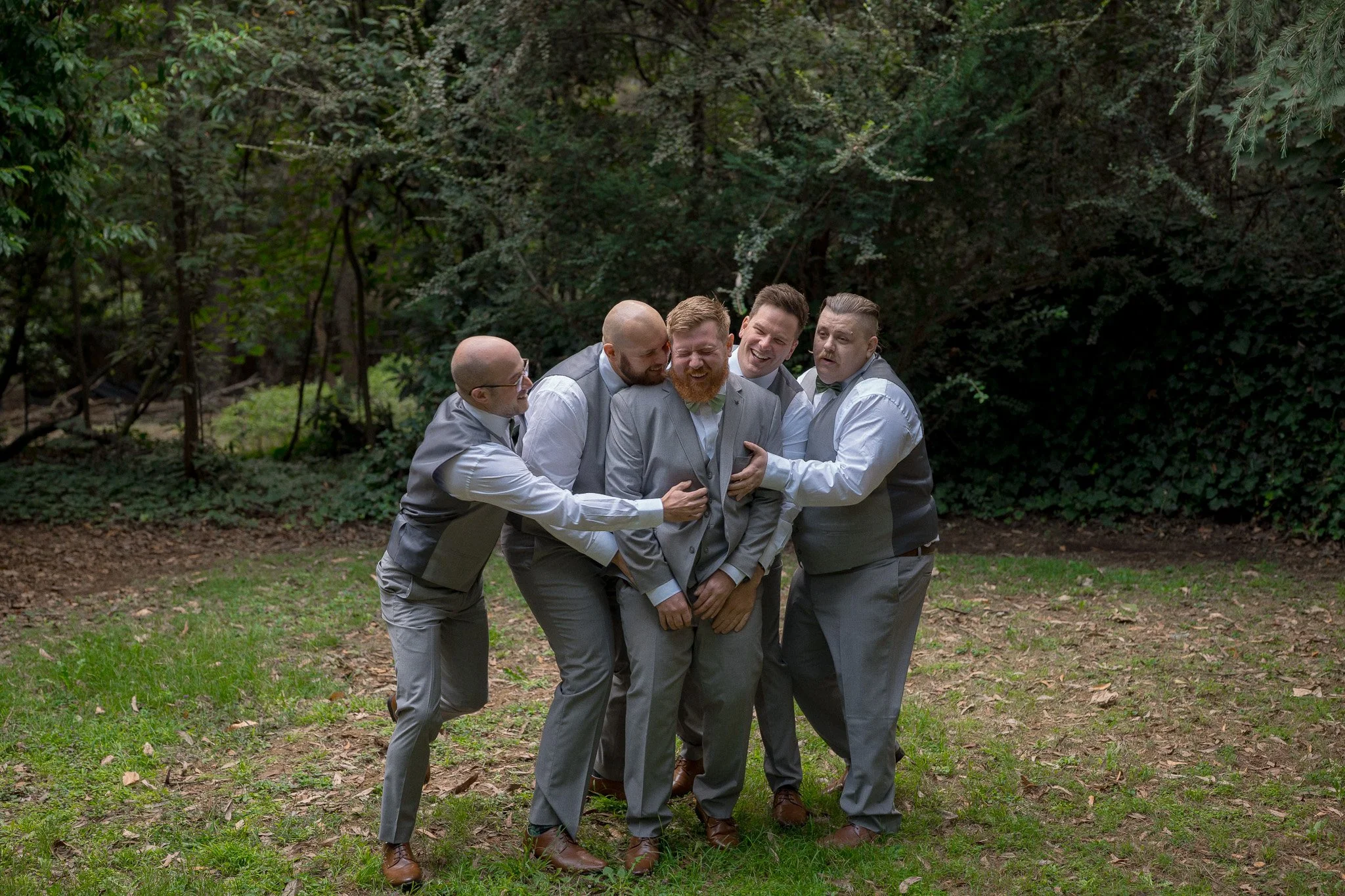 Group of five men in suits enjoying a humorous moment outdoors in a green, wooded area.