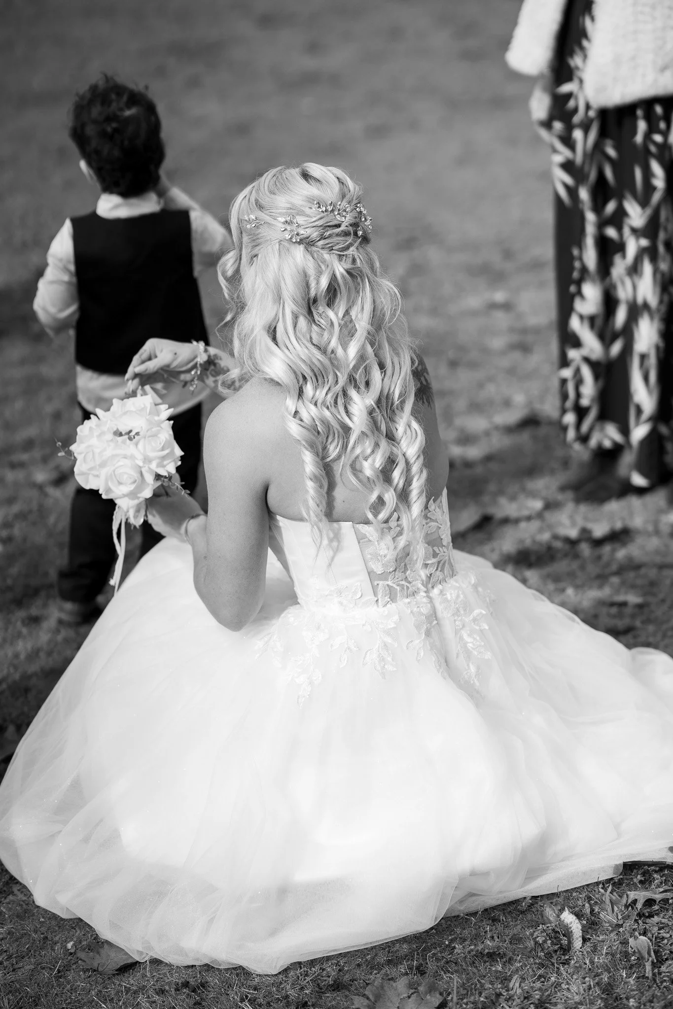 Black and white photo of a bride with long, curly hair wearing a wedding dress and holding a bouquet, standing on grass. A young boy in vest and shirt is in the background.
