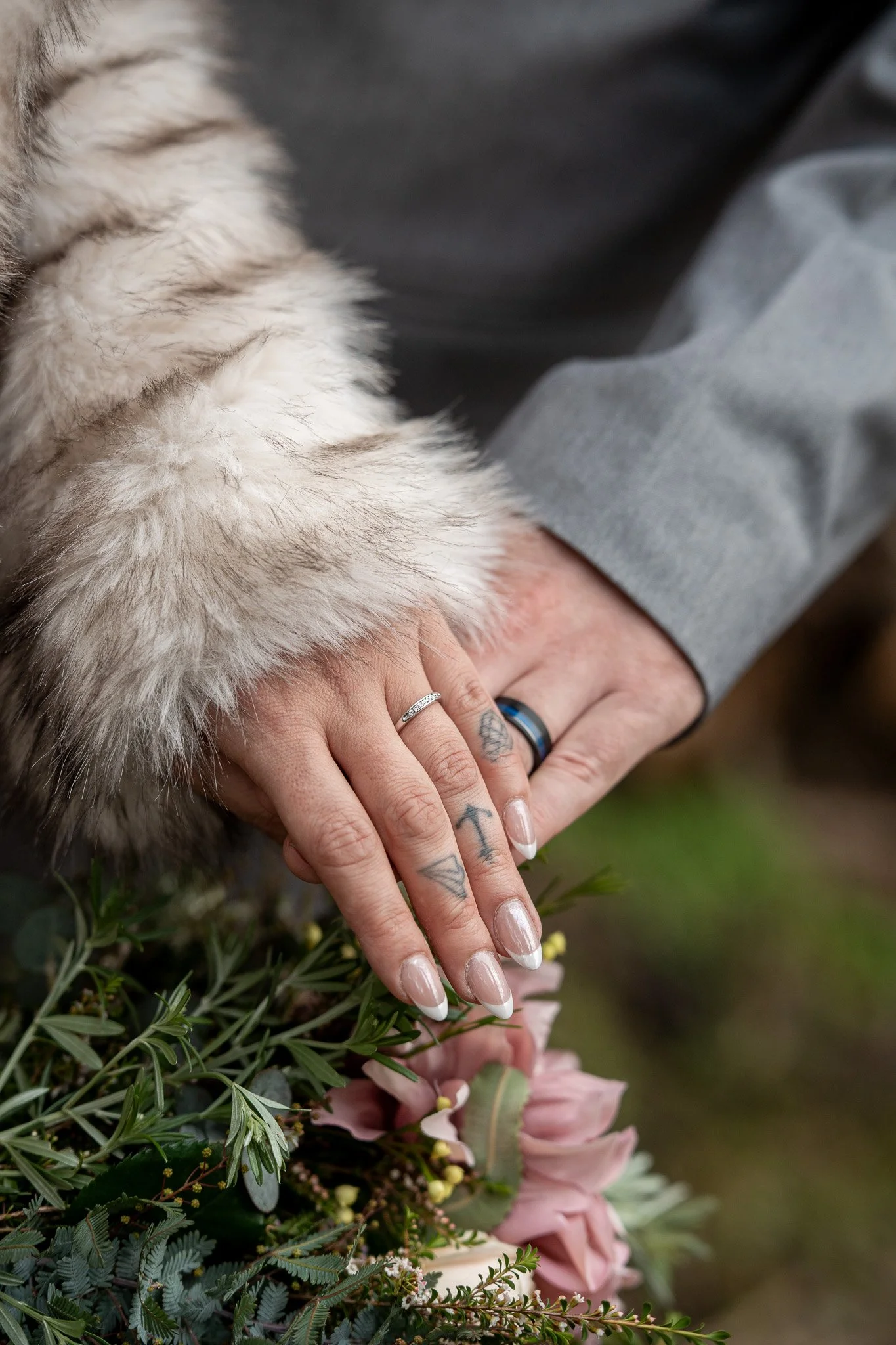 Close-up of a person's hand with tattoos, wearing rings, resting on a bed of assorted flowers and greenery.
