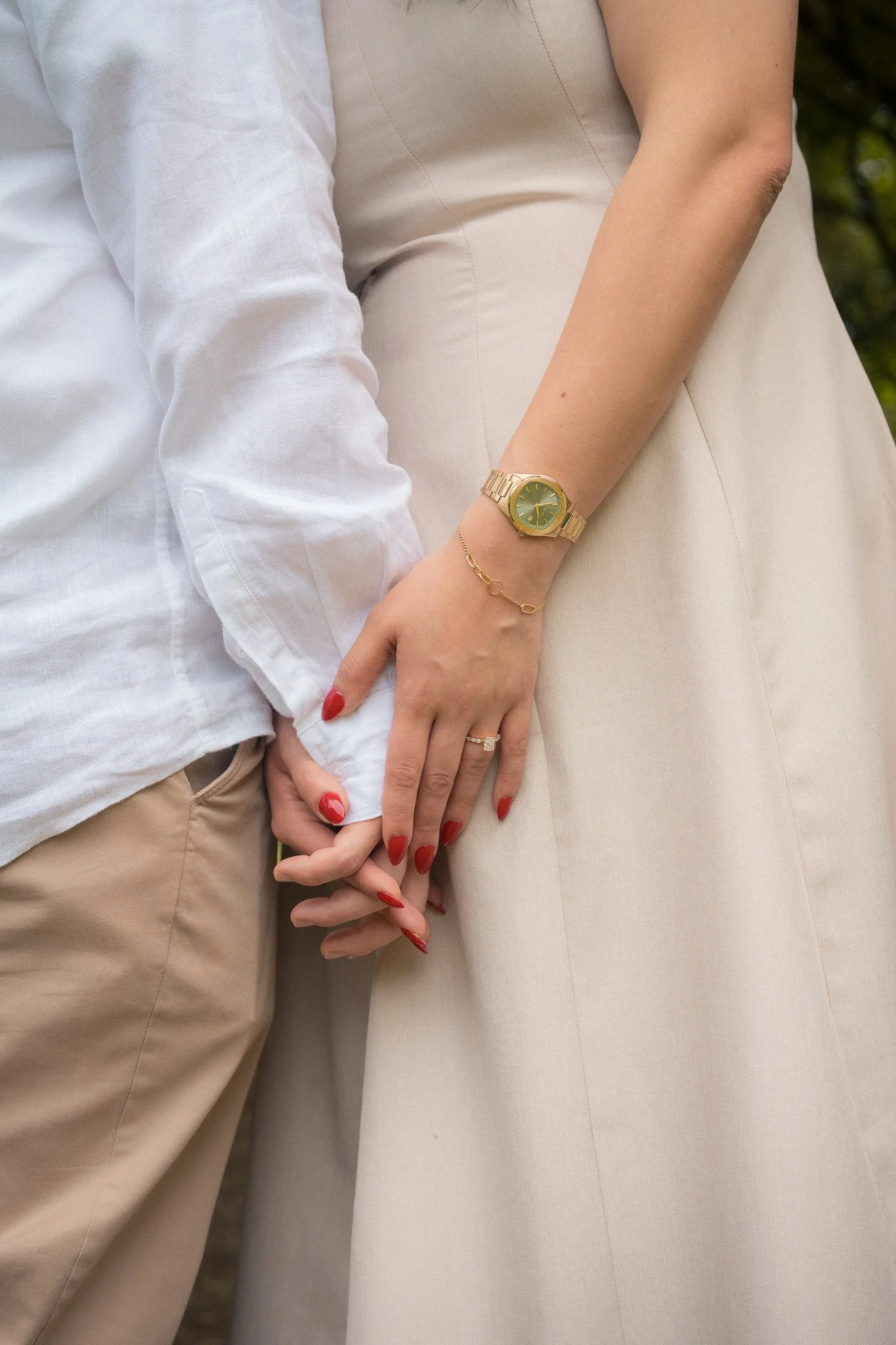 Close-up of a man and woman holding hands, with the woman's hand resting on the man's arm. The woman wears a gold watch, a bracelet, and a diamond ring, with red painted nails. Both are dressed in light-colored clothing.