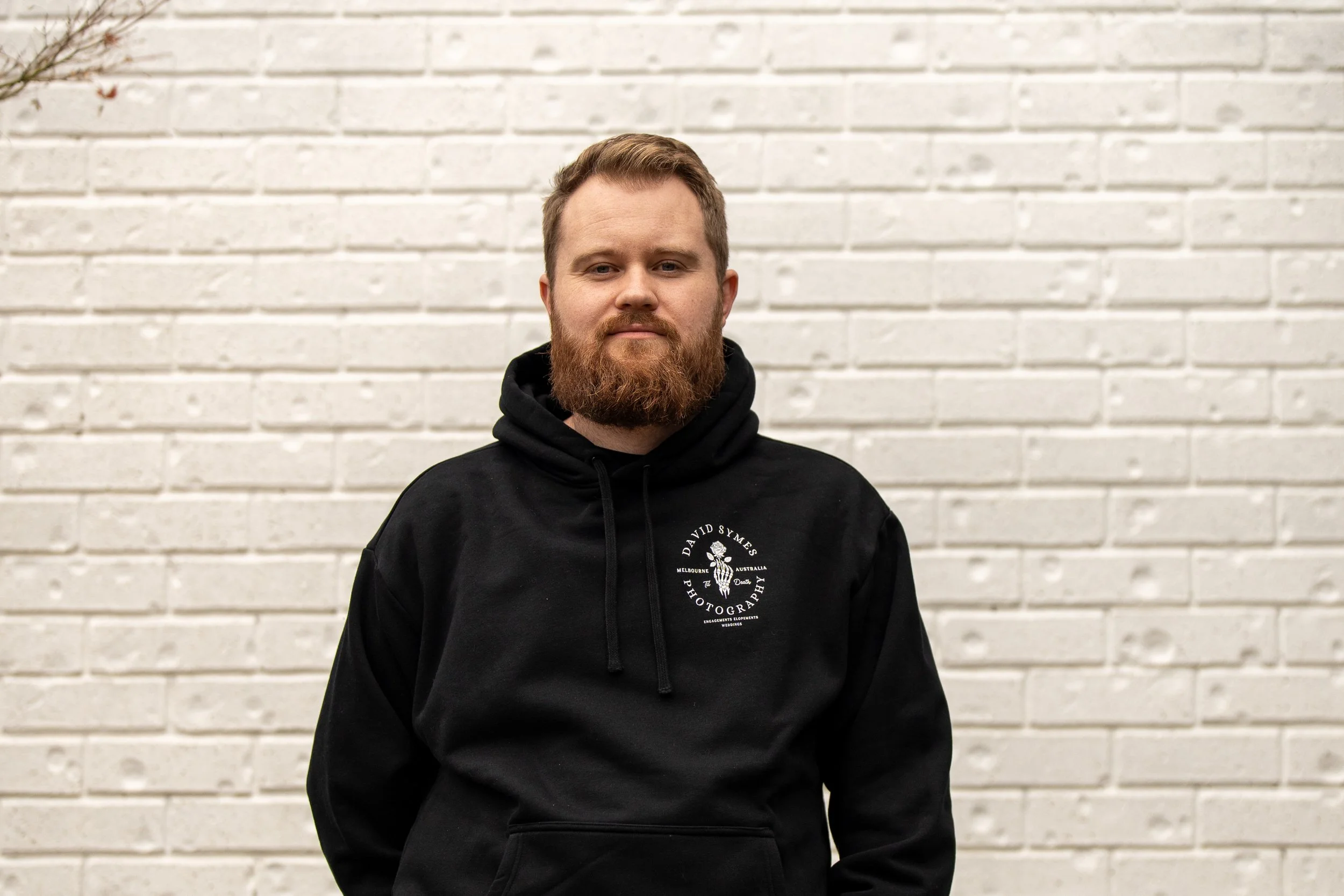 A man with a full beard and short hair stands in front of a white brick wall, wearing a black hoodie.