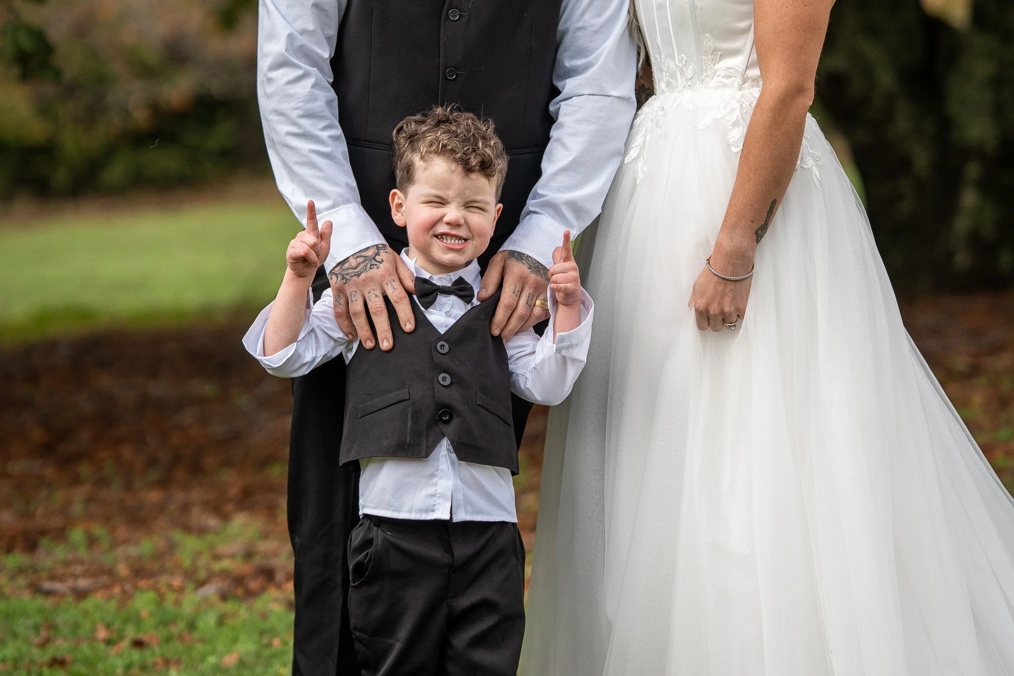 Close-up of a young boy in a tuxedo, making a funny face, with an older man and woman dressed in wedding attire standing behind him, outdoors with green foliage in the background.