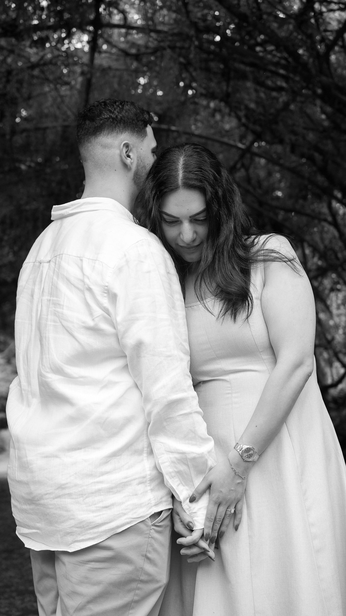 A black and white photo of a couple standing close, holding hands, with the woman looking downward, outdoors with trees in the background.