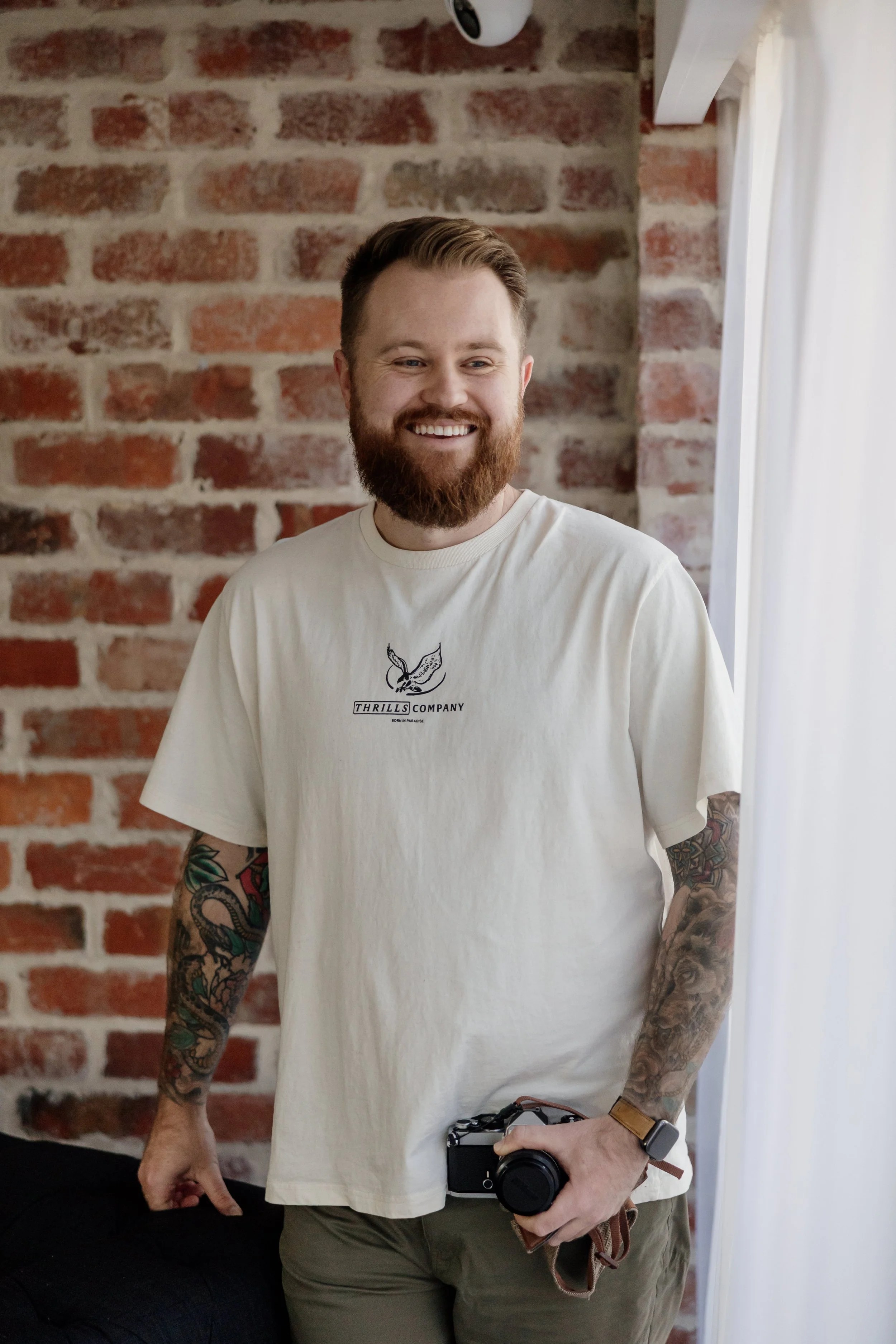 A man with a beard and tattoos on both arms is smiling while holding a camera and standing in front of a brick wall with white curtains on the right side.