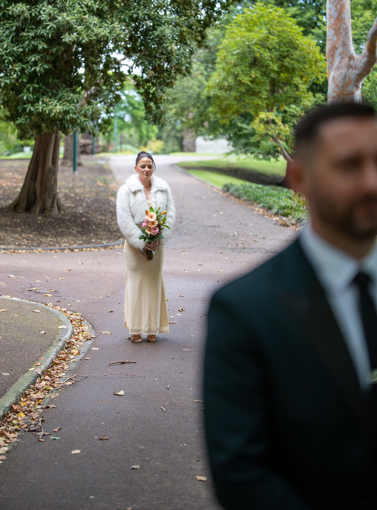 A woman in a white faux fur jacket and a long cream dress holding a bouquet of flowers, standing on a park path, with a man in a dark suit blurred in the foreground.