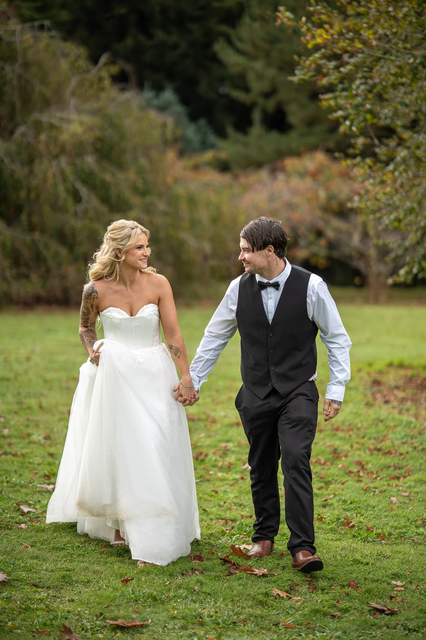 A bride and groom holding hands while walking outdoors in a grassy area with trees and autumn leaves in the background.