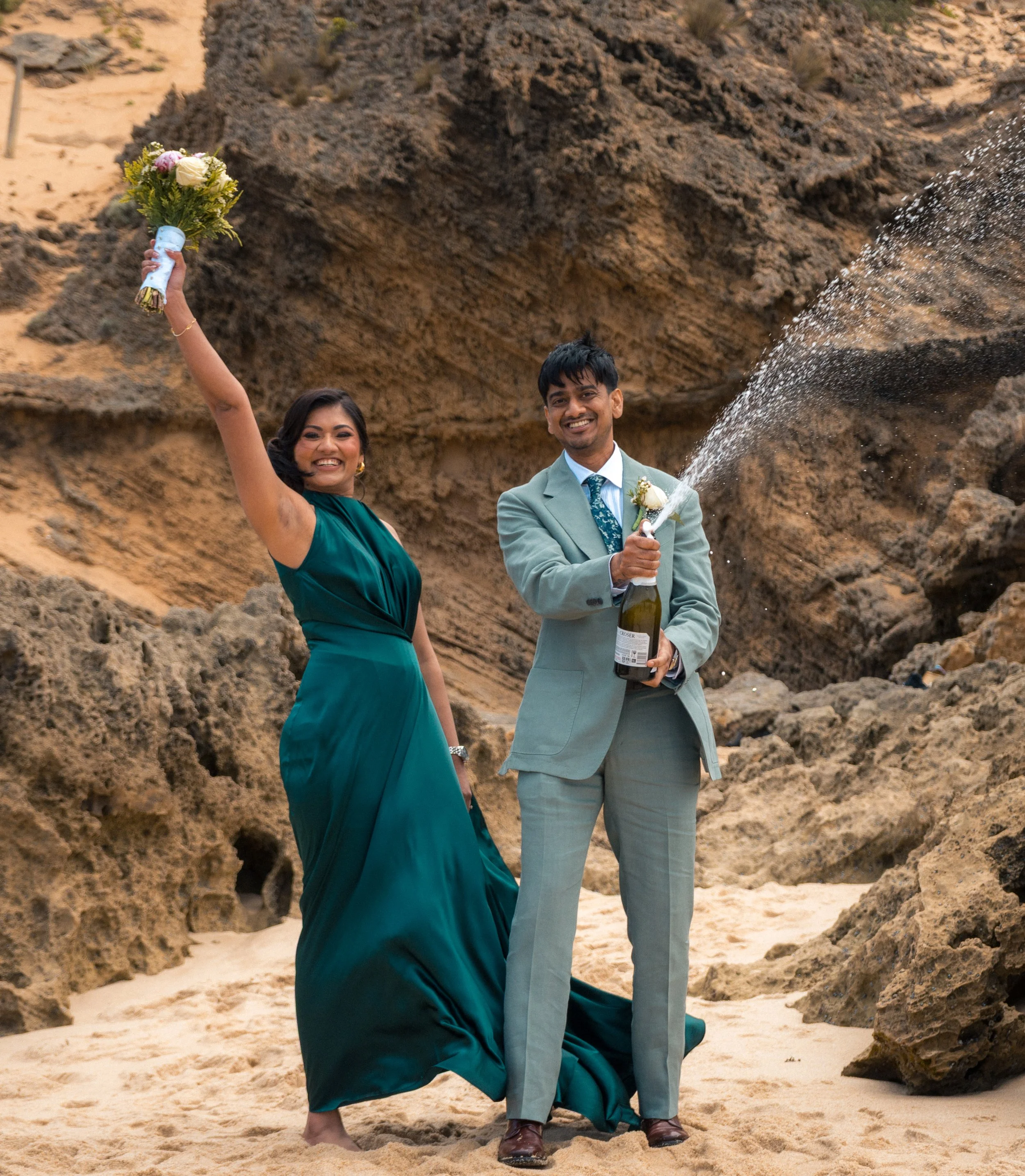 A woman in a teal dress and a man in a gray suit celebrating on a beach with rocky cliffs, holding a bouquet and popping champagne, smiling.
