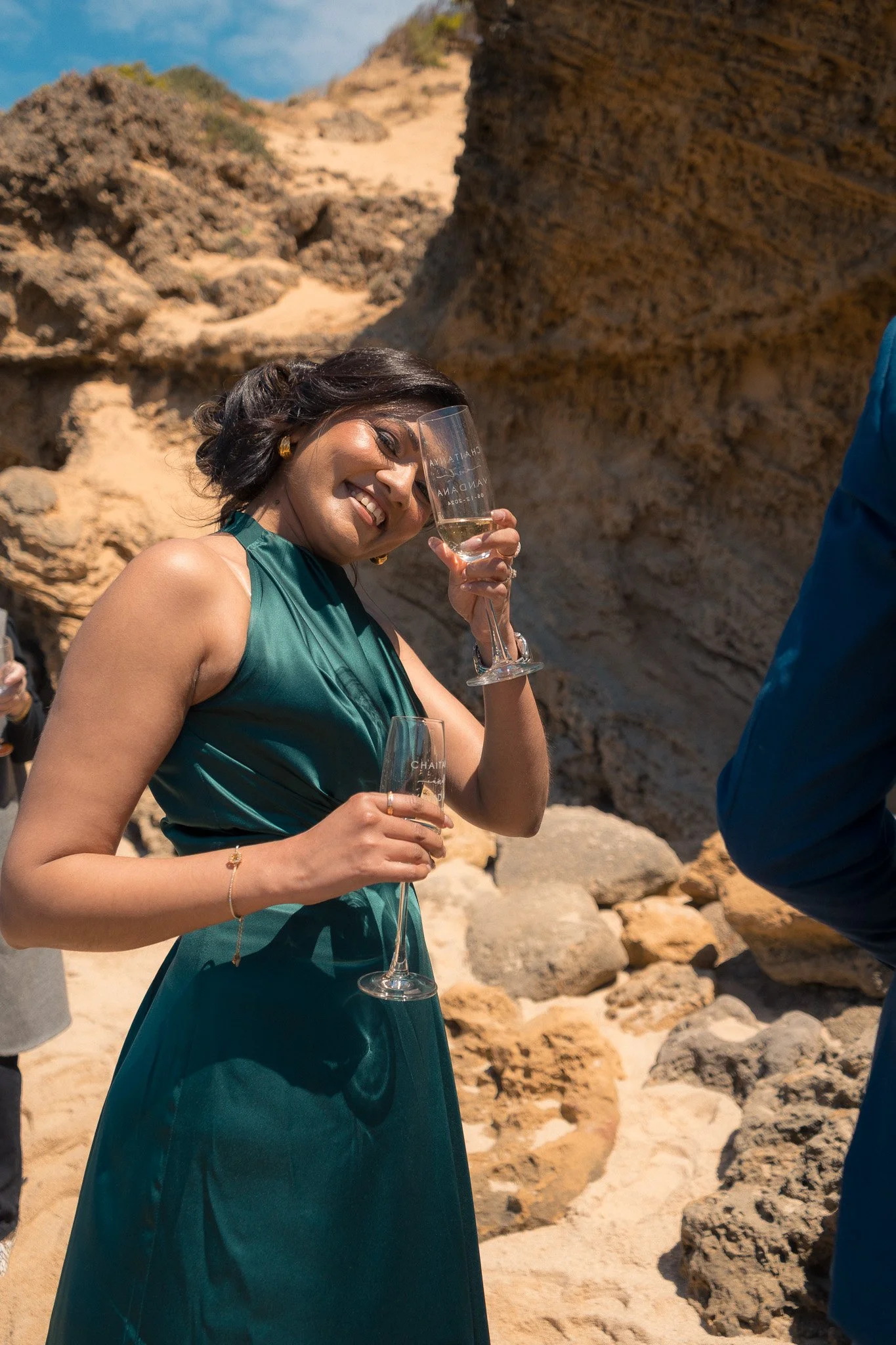 A woman in a green dress smiling and holding a champagne flute, standing outdoors on a rocky beach with cliffs in the background.