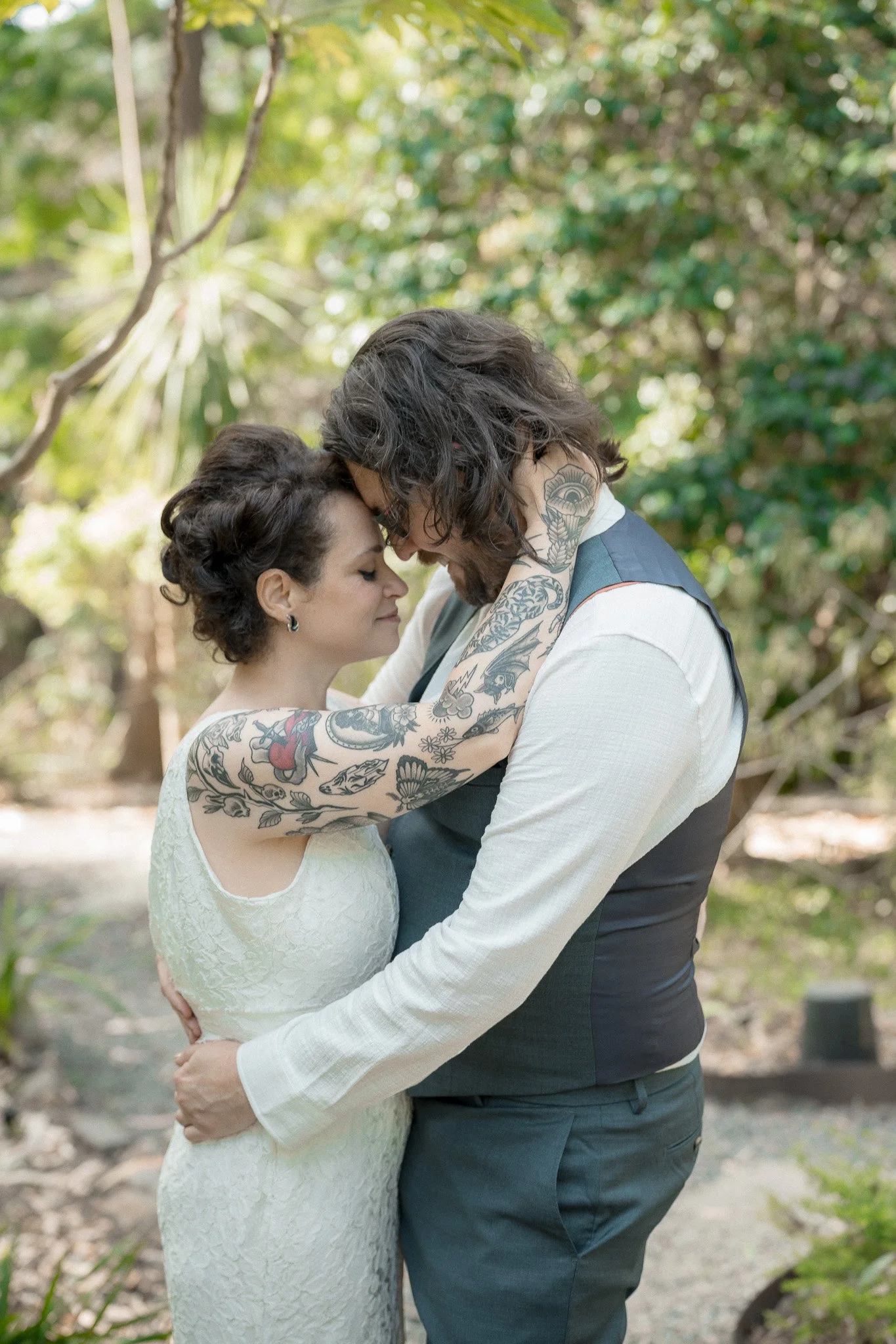 A couple embracing outdoors, with foreheads touching, eyes closed, surrounded by greenery.