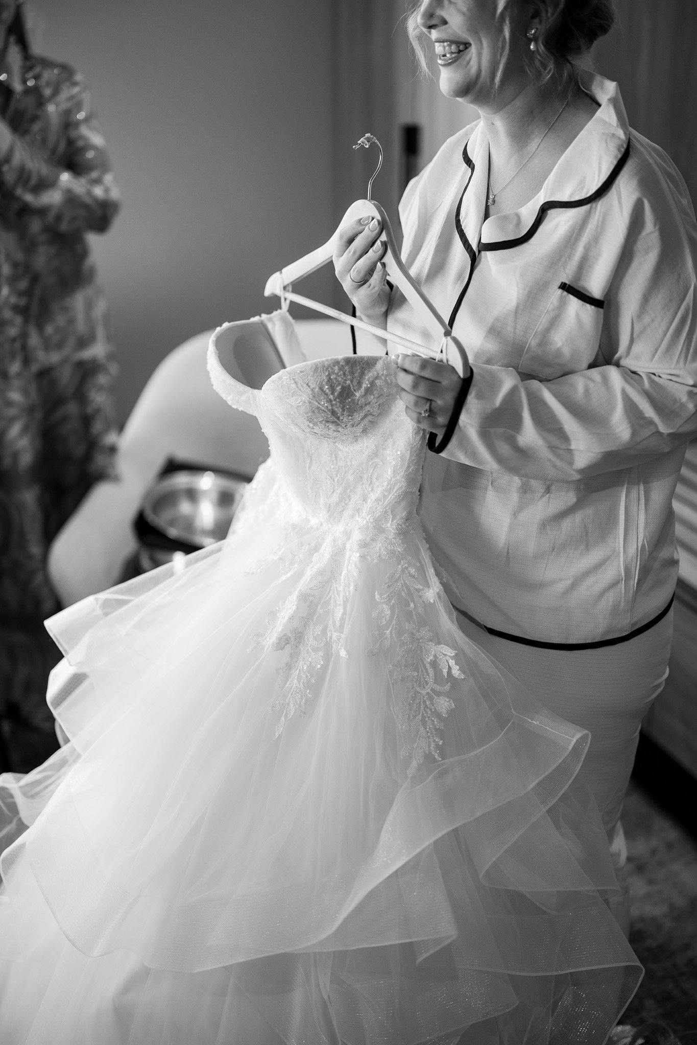 A woman in pajamas holds a wedding dress on a hanger and smiles.